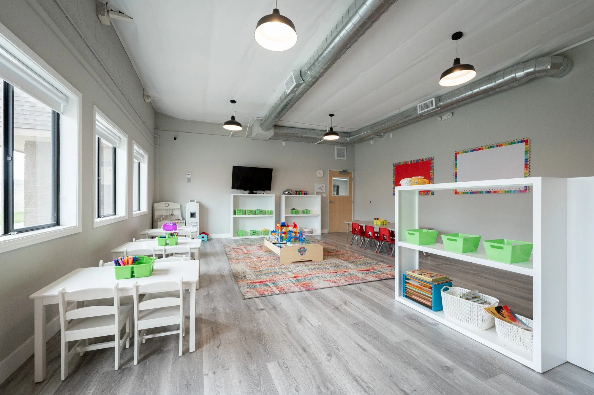 Bright classroom with large windows, white tables with green and purple containers, white chairs, a colorful rug with children's toys, shelves with green bins, and colorful bulletin boards on the walls. A wall-mounted TV and a door are visible.