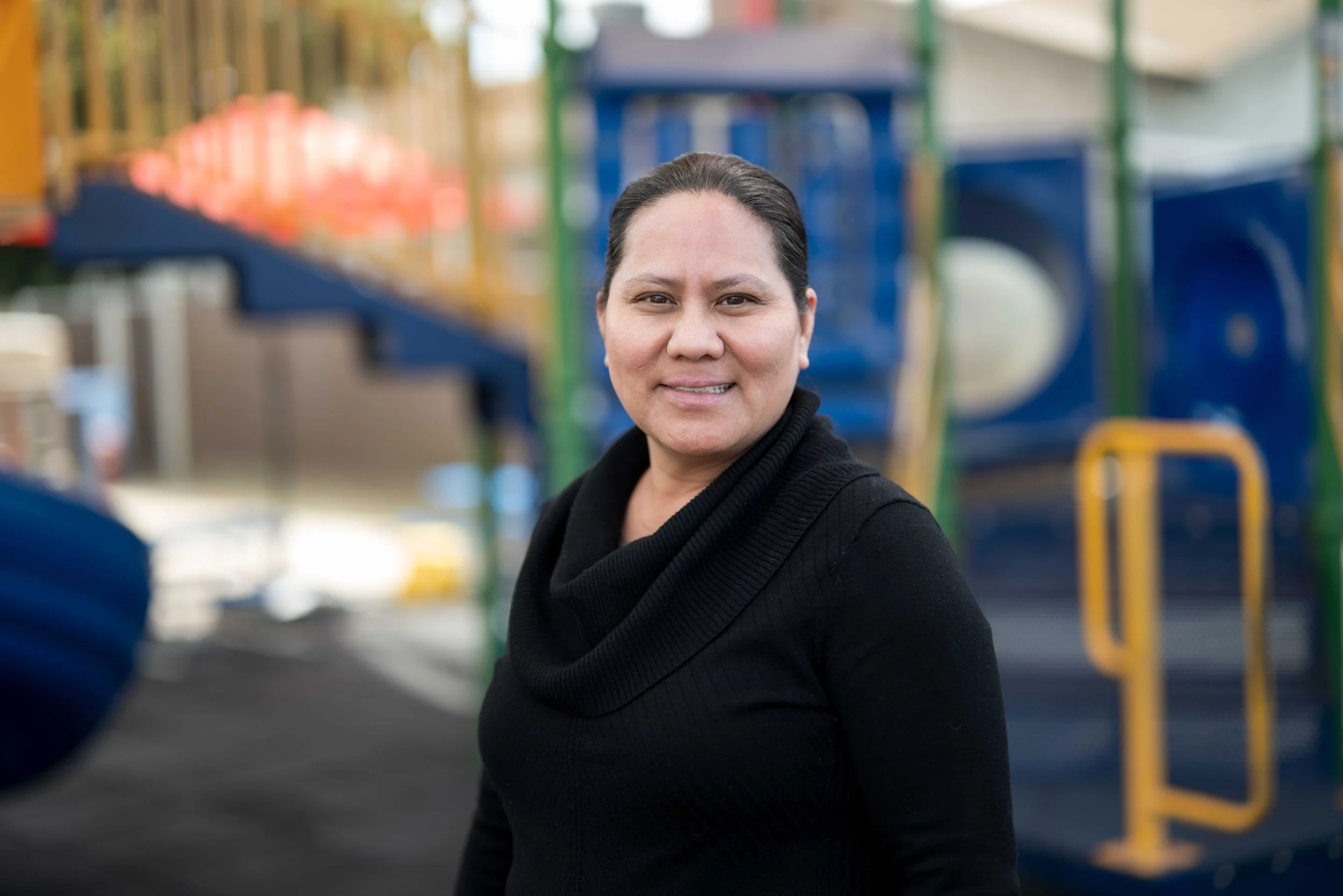A woman with dark hair pulled back, wearing a black top, is smiling outdoors with a colorful playground in the background.