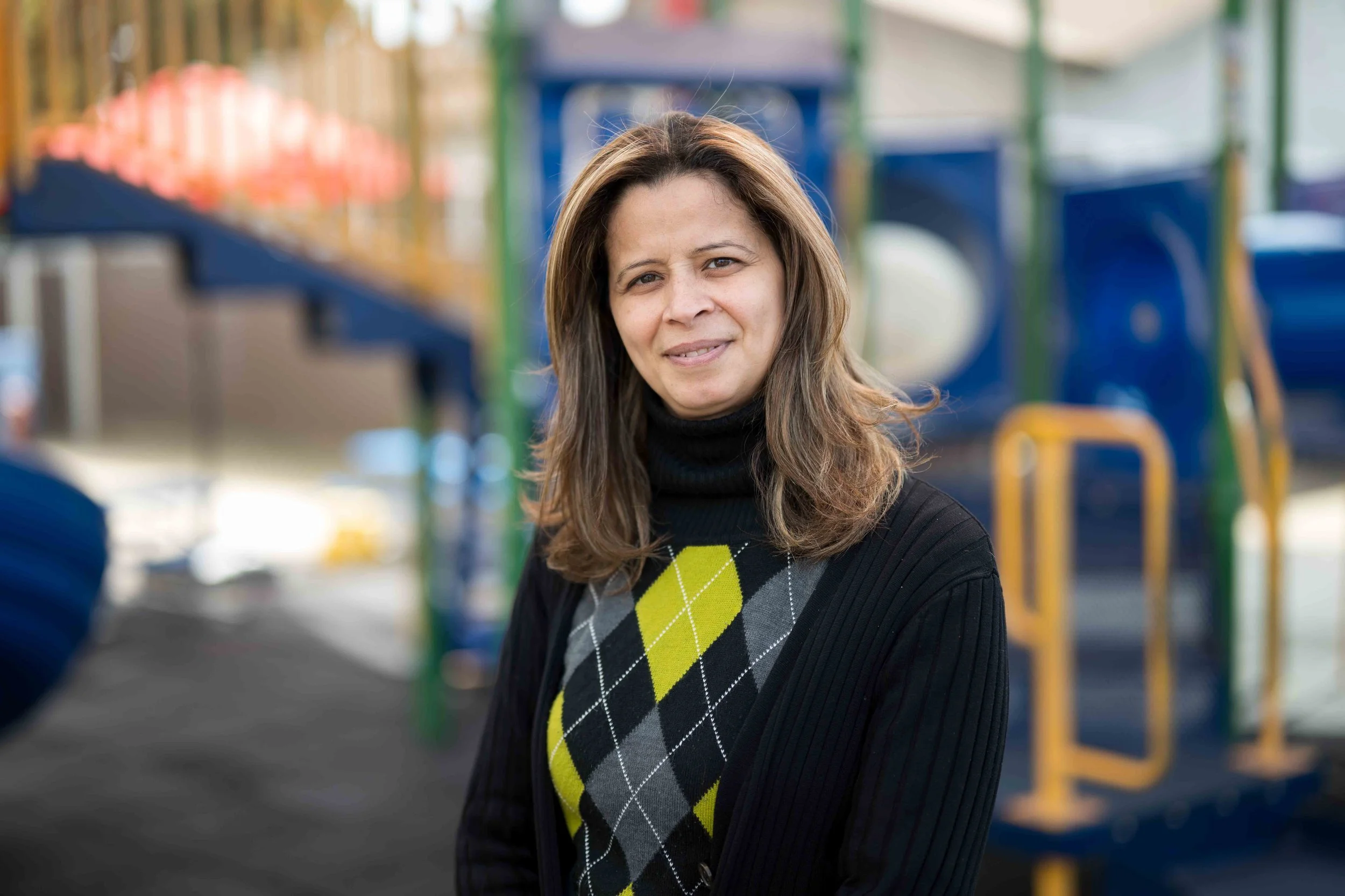 A woman with long brown hair wearing a black turtleneck sweater and an argyle patterned vest poses outdoors at a playground.