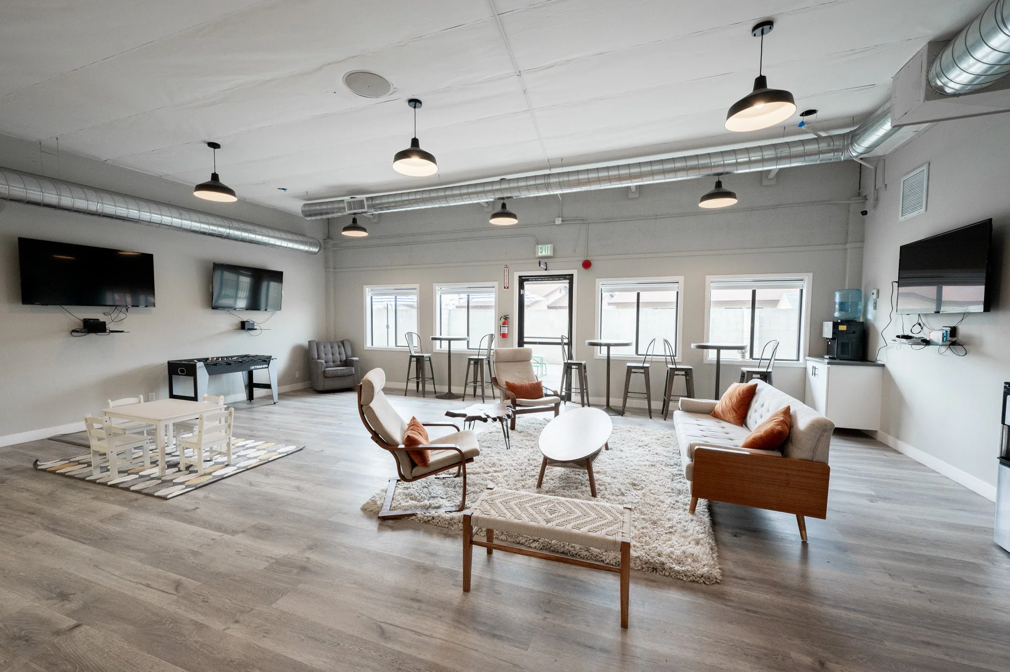 A spacious lounge area with multiple seating options including armchairs and sofas, a large shaggy rug, a foosball table, three wall-mounted TVs, and a drinking water dispenser, with large windows, modern ceiling lights, and exposed ductwork.