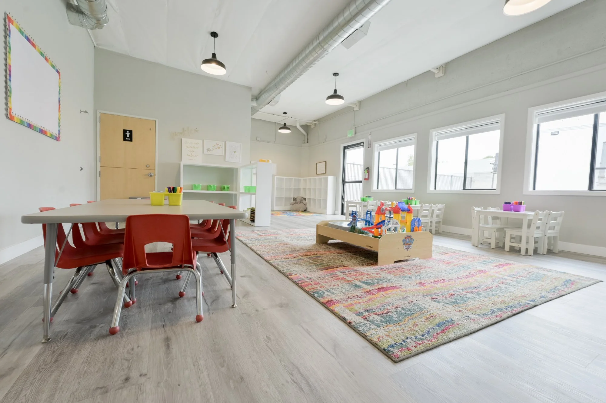 Indoor children's play and learning area with tables, chairs, and toys, near large windows allowing natural light, with colorful rug and wall decorations.