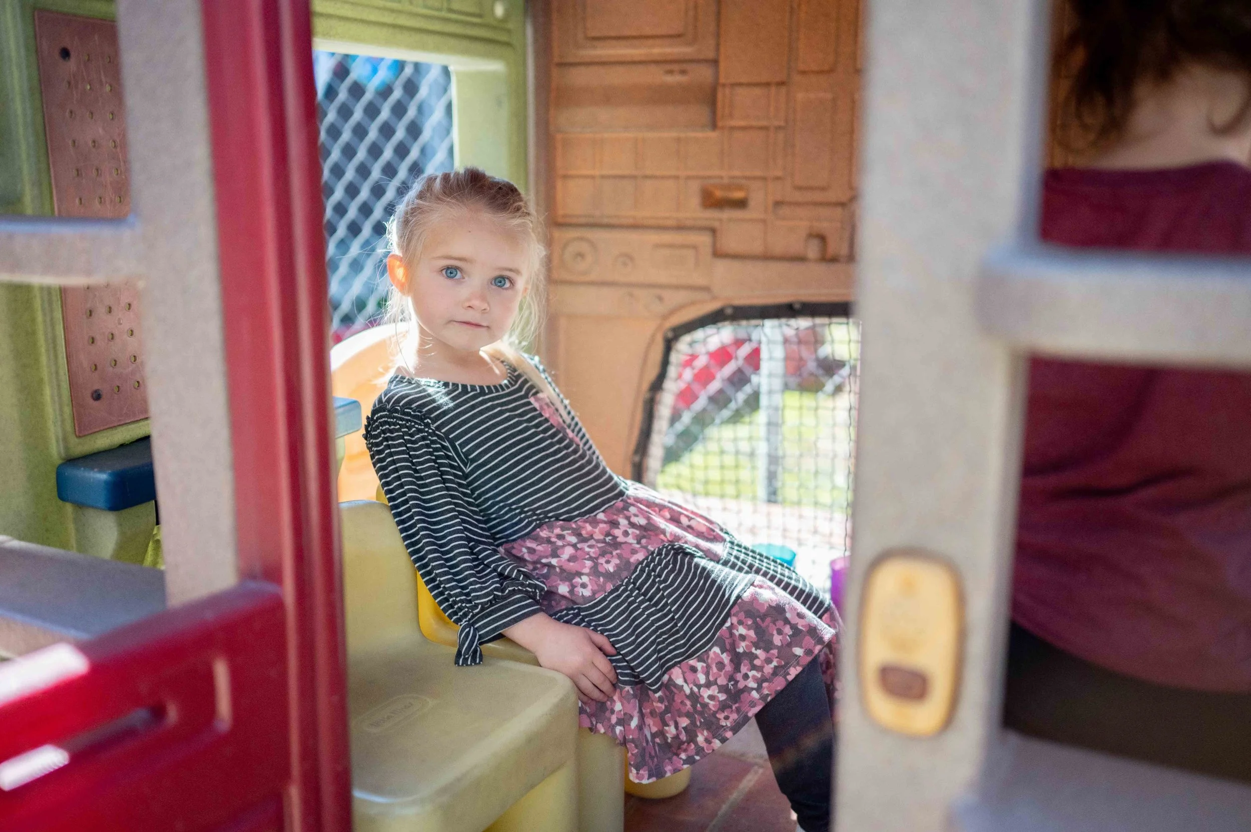Young girl with curly hair and bright blue eyes sitting on a yellow seat inside a playhouse made of wood and plastic. The playhouse has colorful walls and a window with a mesh screen in the background.