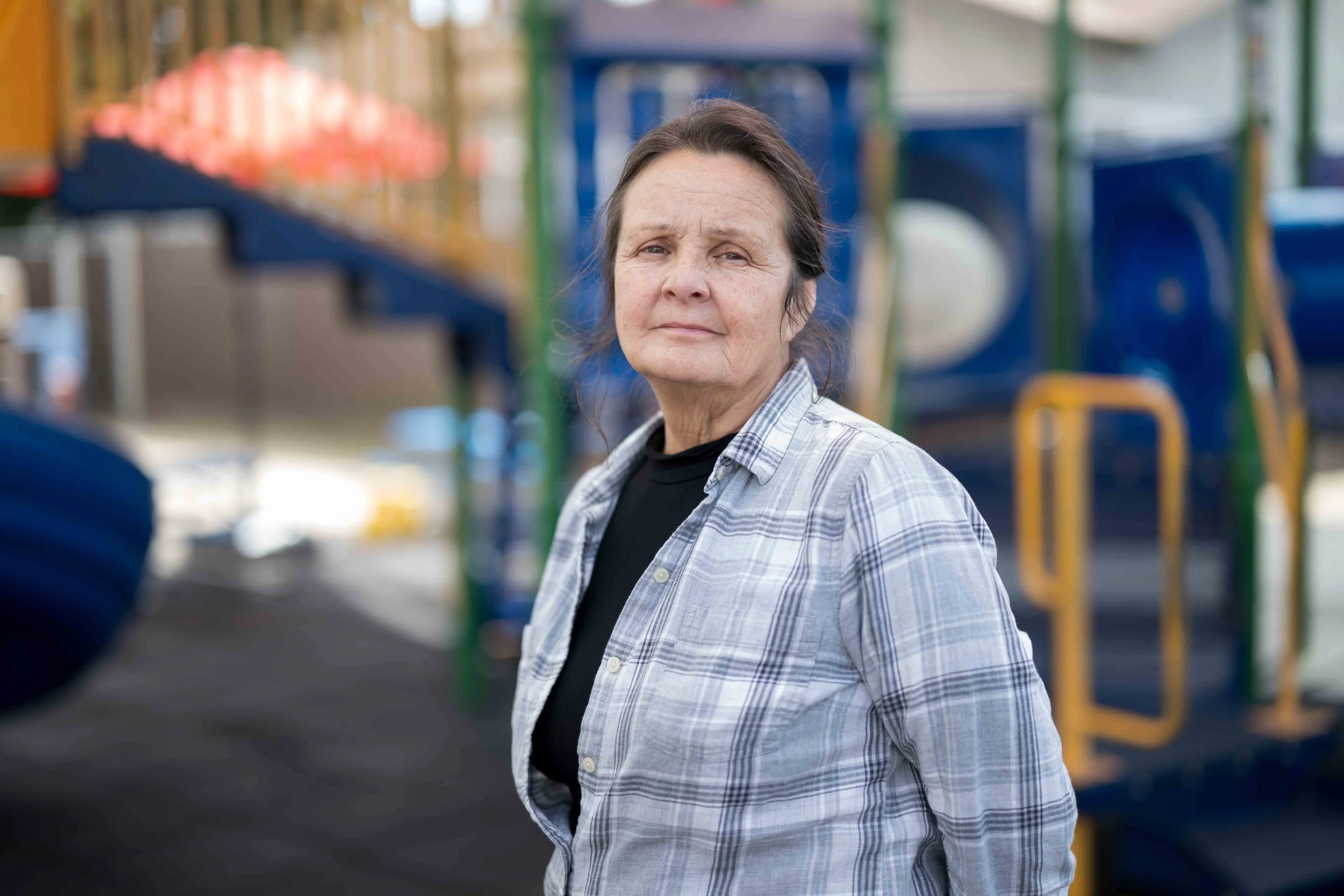 A middle-aged woman with dark hair and a neutral expression, wearing a gray plaid shirt over a black top, standing in front of a colorful playground structure.