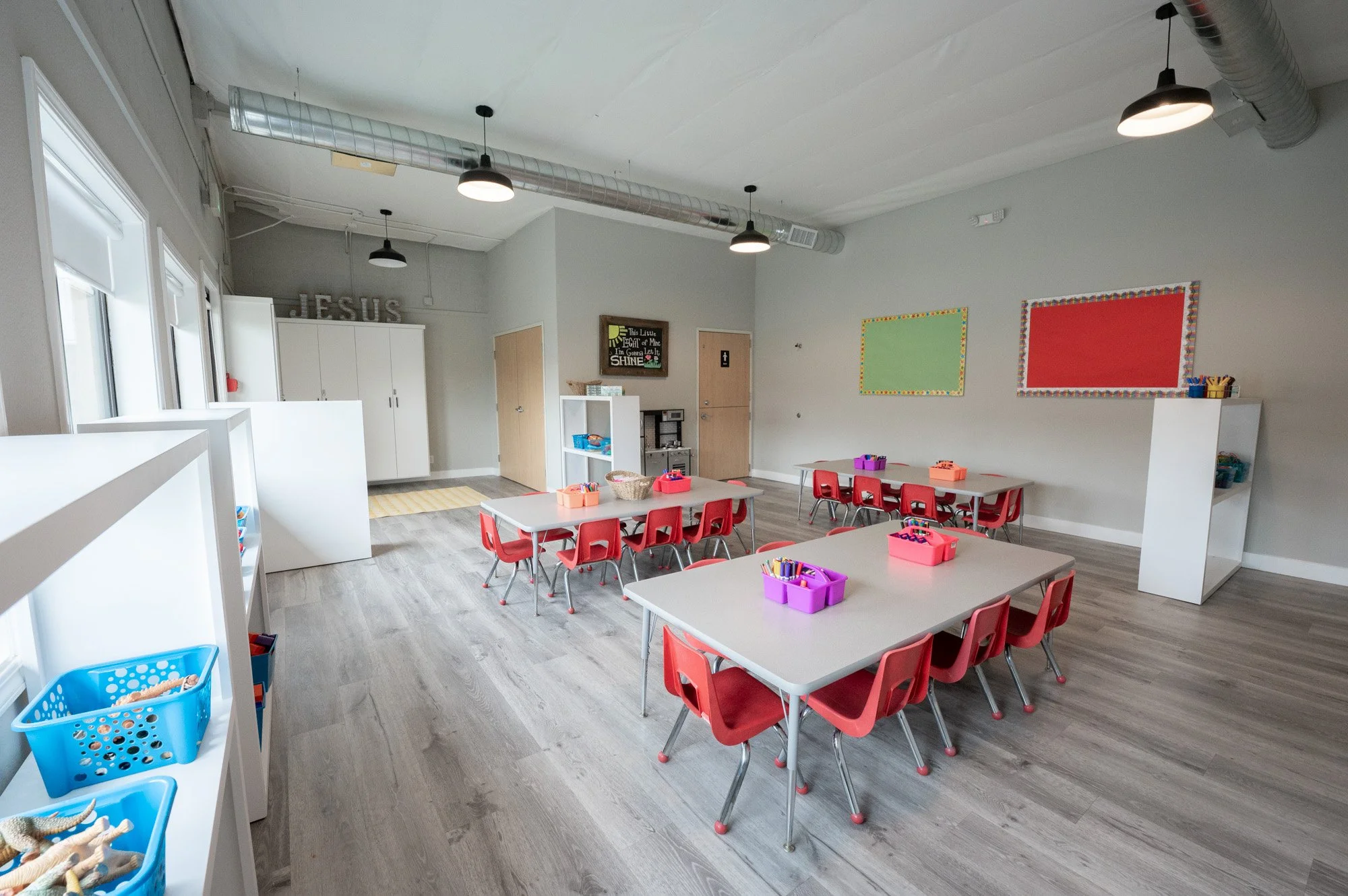 A spacious classroom with three tables surrounded by red chairs, each table has baskets filled with art supplies like markers and crayons. There are white bookshelves on the side with additional supplies, and windows along the left wall allowing natu