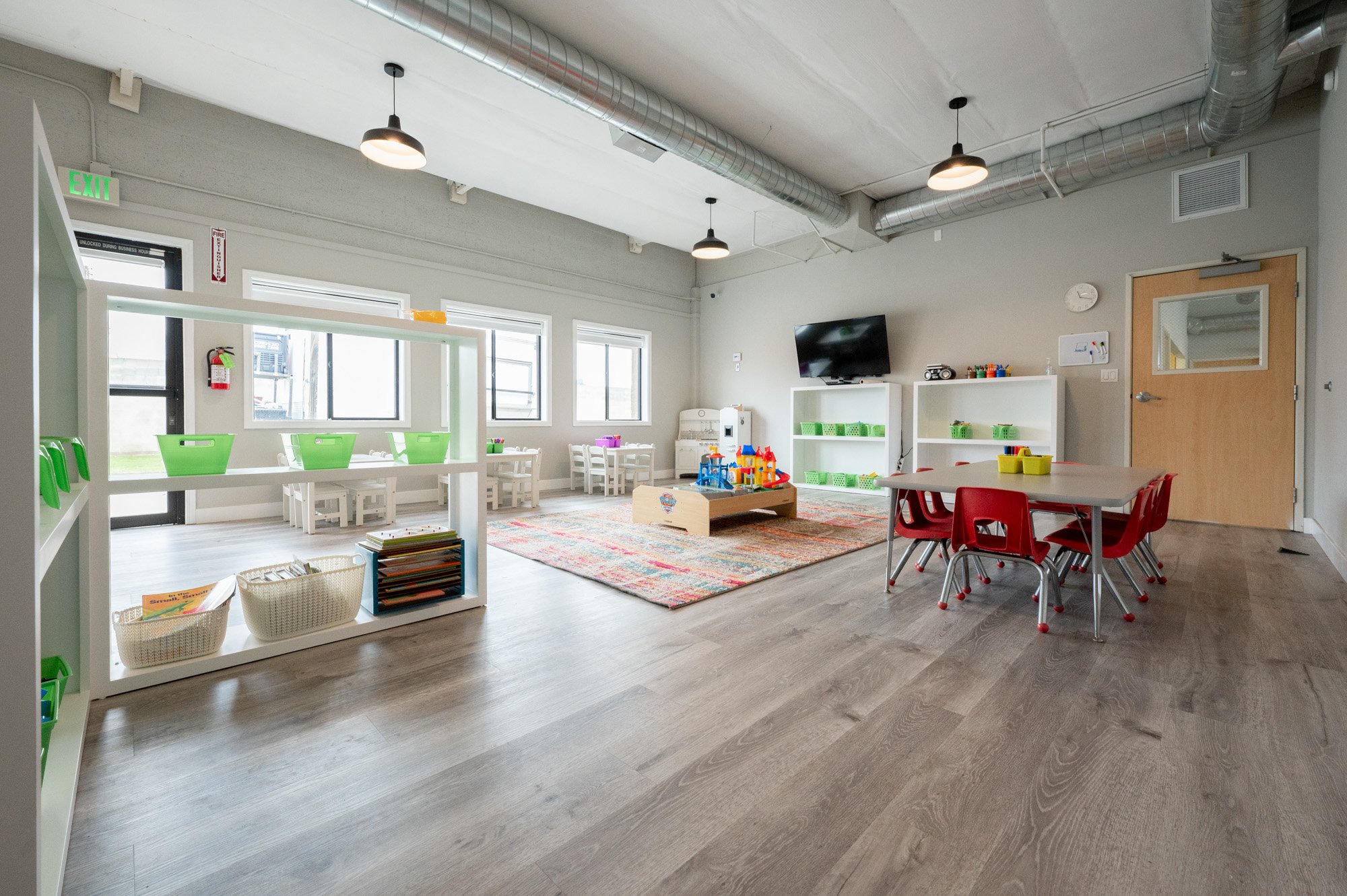Children's classroom with tables, chairs, and toys, bright and organized, wall-mounted TV, windows, and ceiling lights.