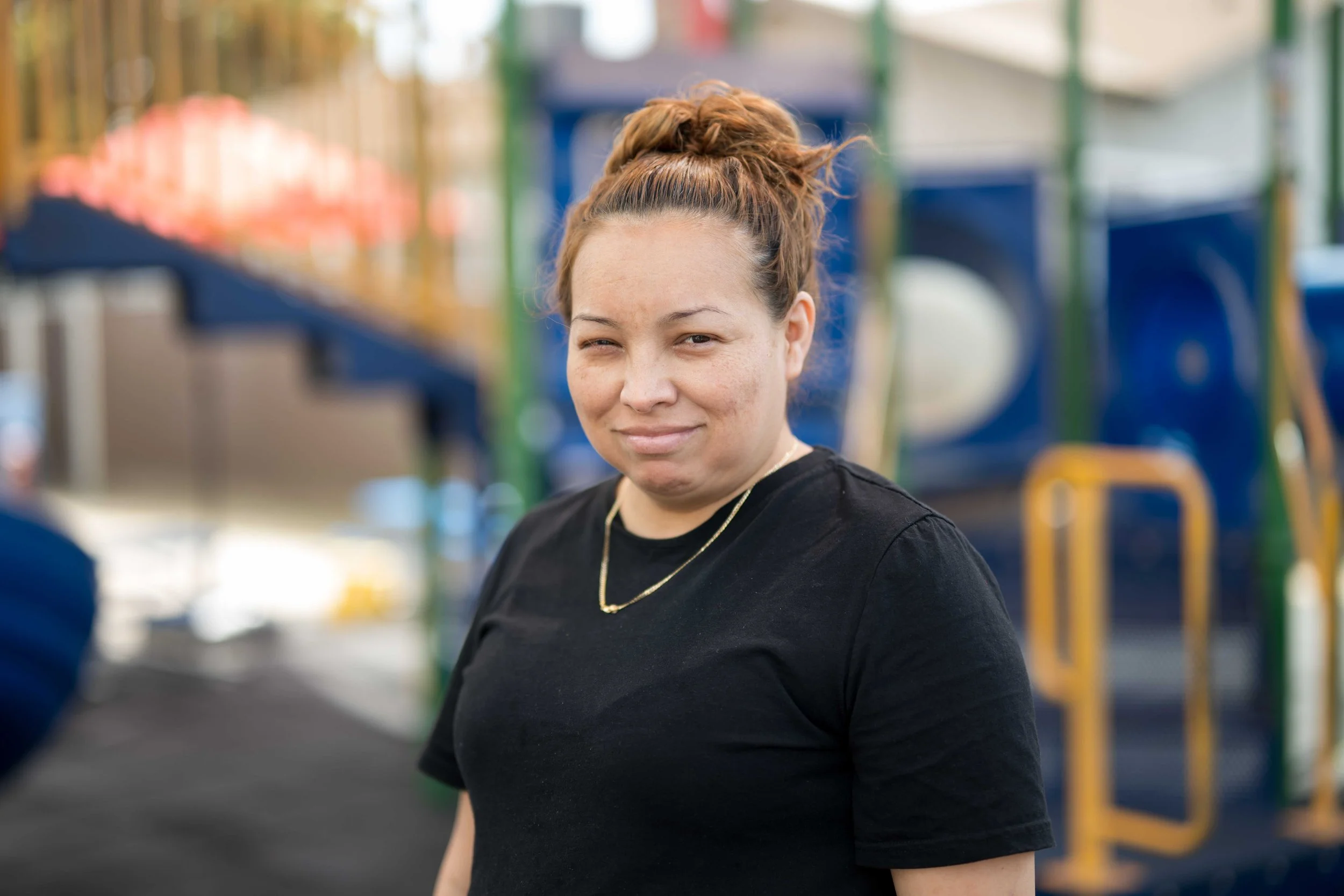 A woman with brown hair styled in a high bun, wearing a black shirt and a gold necklace, smiling at camera in an outdoor playground setting.