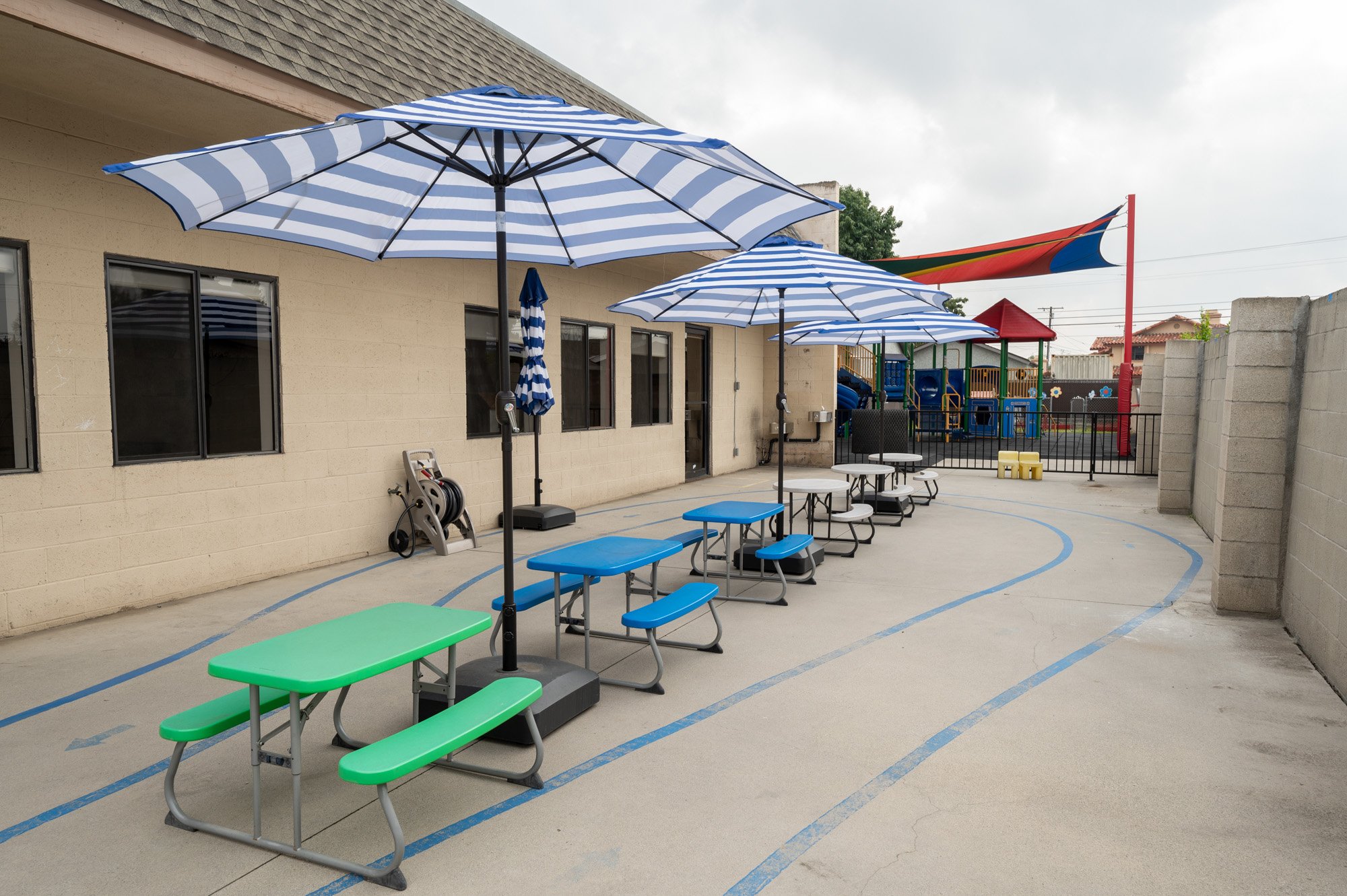 School playground with blue and white striped umbrellas over picnic tables, colorful play structure with slides and climbing areas, concrete walls, and a red, yellow, blue, and green shade sail, on a cloudy day.