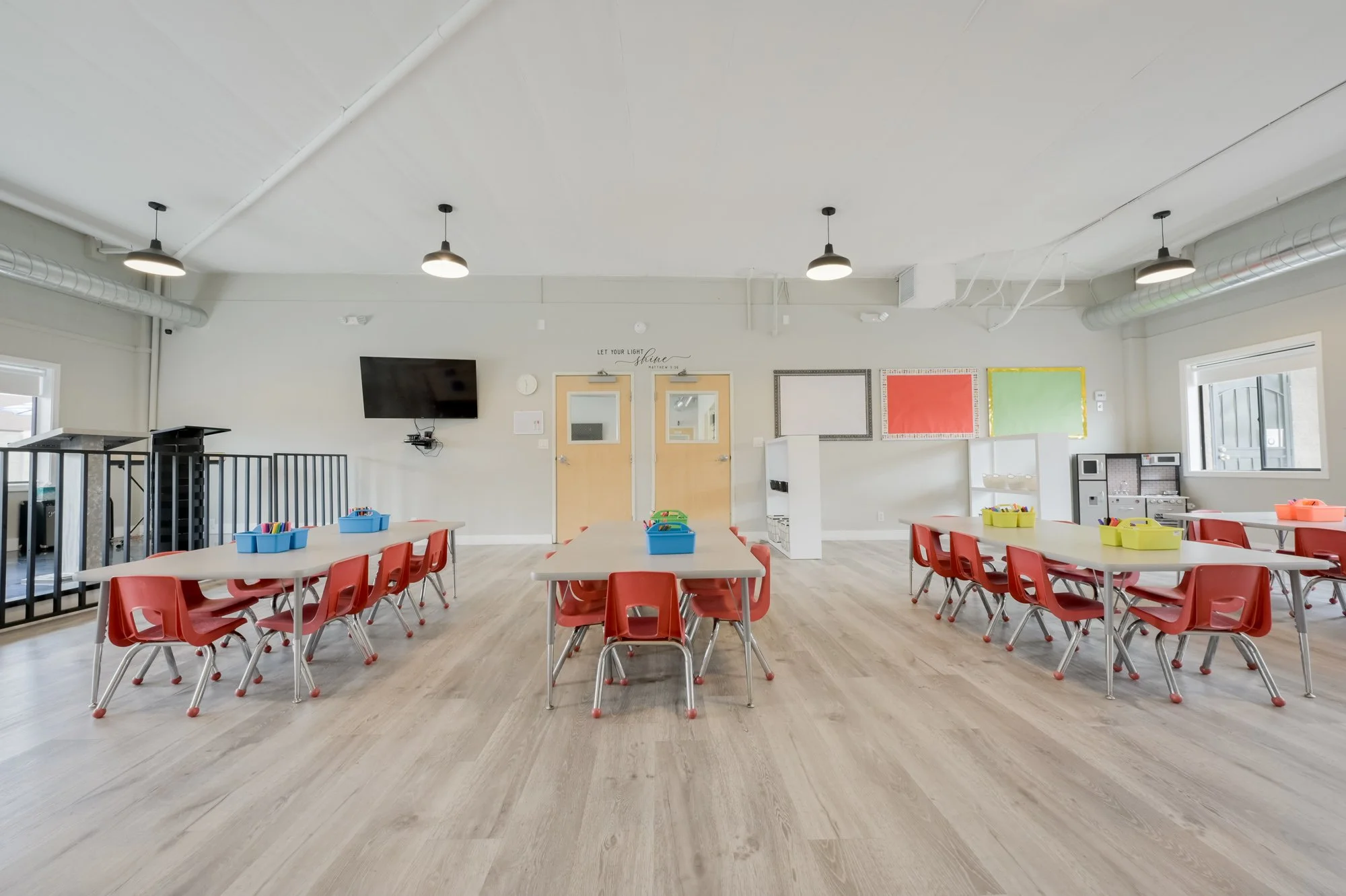 A brightly lit classroom with several rectangular tables surrounded by red chairs. There are small blue and yellow bins on the tables. The walls have colorful bulletin boards. A black television, wall clock, and framed artwork are visible on the back