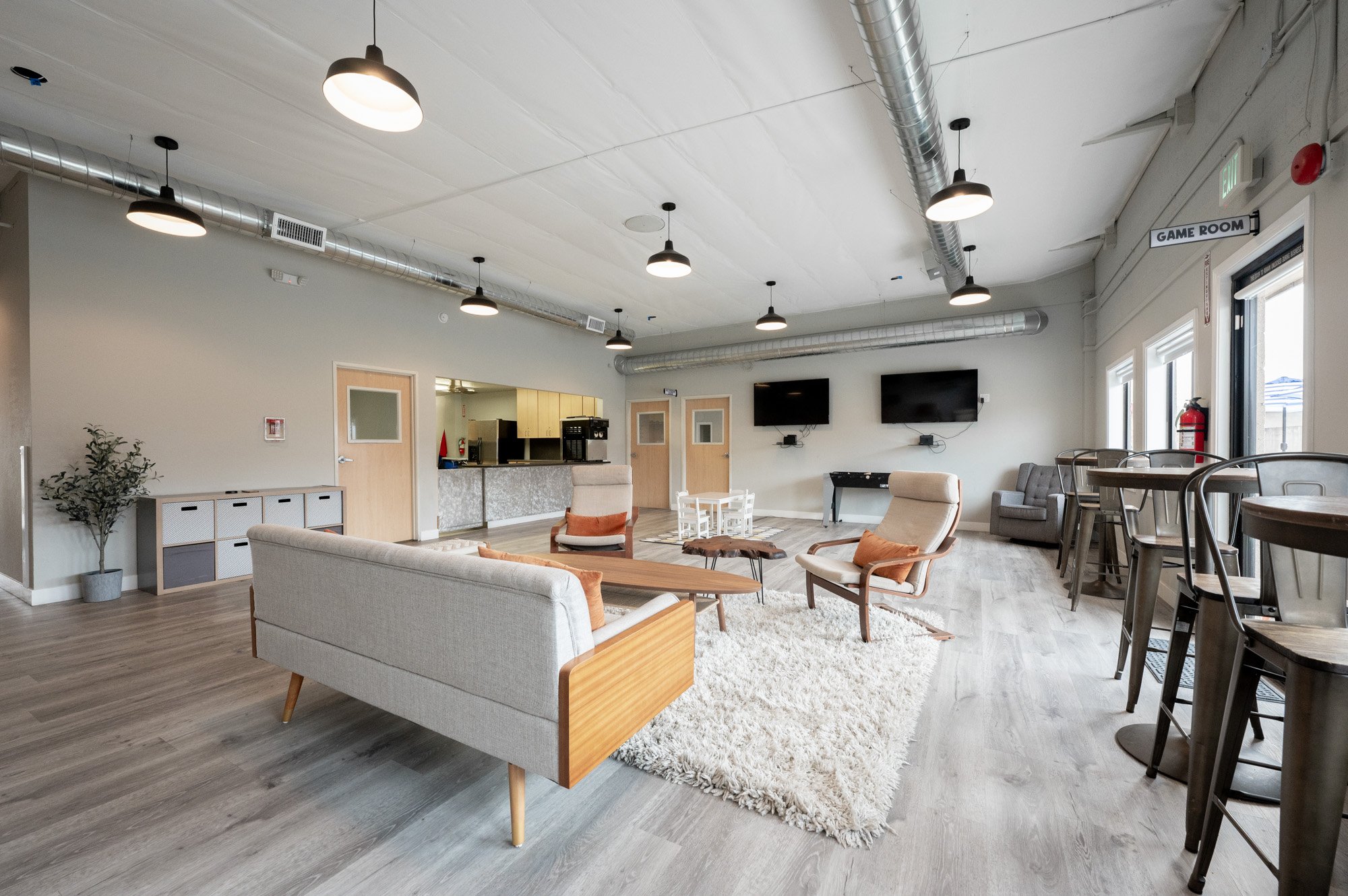 A spacious, modern lounge area with a sofa, armchairs, a wooden coffee table, TV screens on the wall, a kitchenette, and a sign indicating a game room, decorated with sleek lighting and metal ductwork.