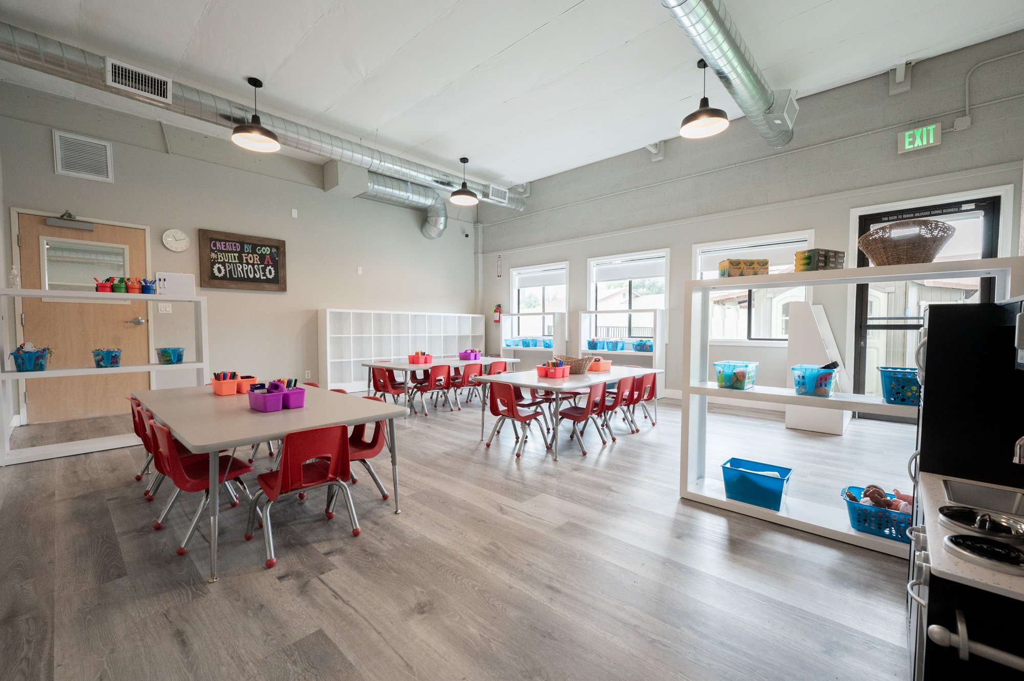 A bright classroom with small red chairs and tables, white shelves with colorful bins, and large windows allowing natural light, with air ducts and modern ceiling lights.