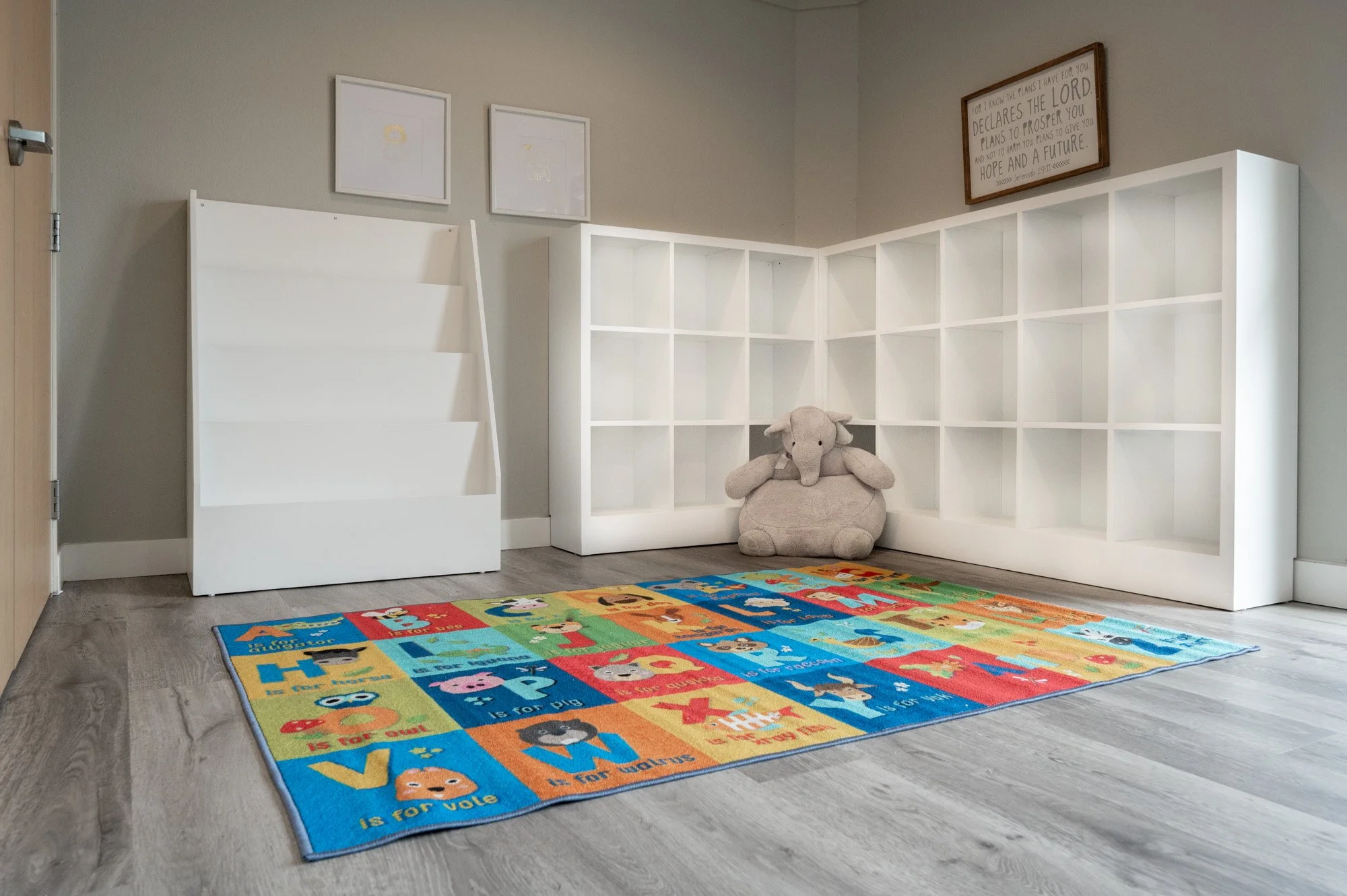 Nursery corner with white cubed bookshelf, stuffed elephant toy, colorful alphabet rug, artwork on the walls, and a framed quote with Christian message.