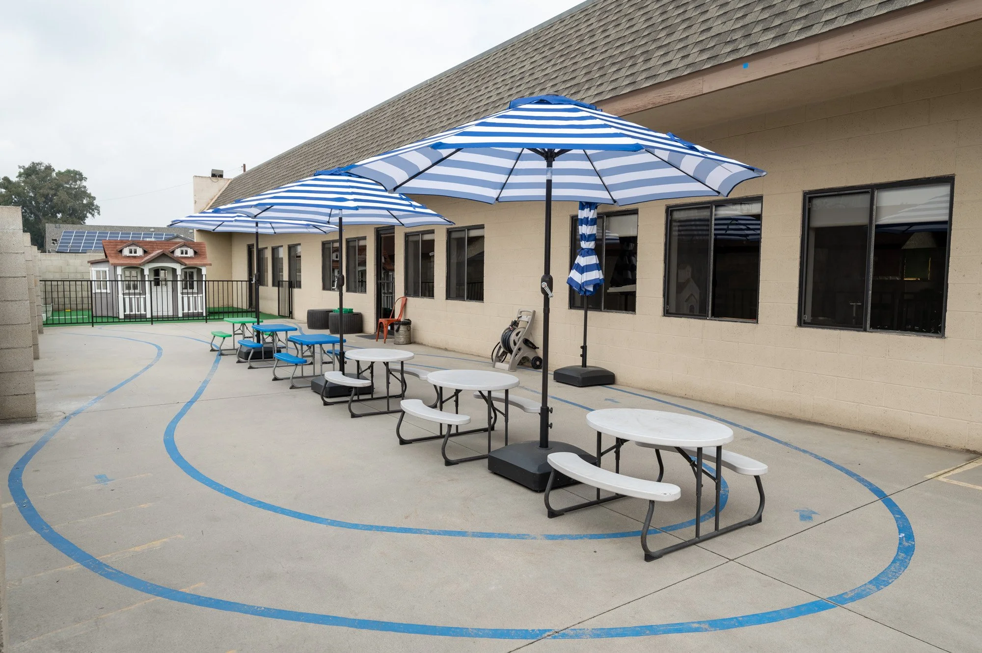 Outdoor school playground with three blue and white striped umbrellas shading picnic tables, a small playhouse in the background, and a concrete surface with blue painted lines.