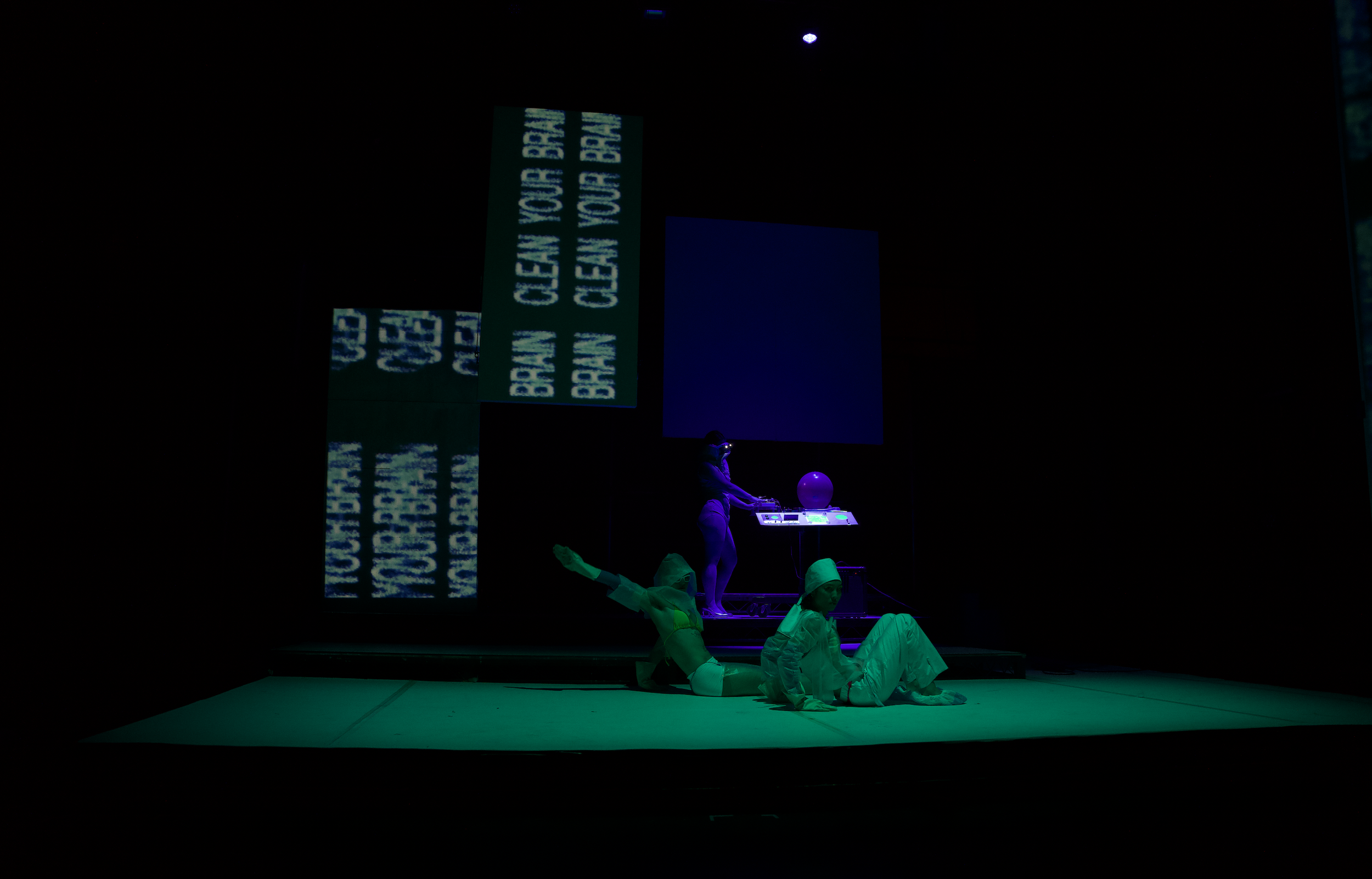 A dark stage with blue lighting displaying text and performers. The text reads: 'TOUCH MEEN YOUR BR...' (partially visible). Performers are dressed in colorful costumes, some lying on the stage, others standing or operating equipment.