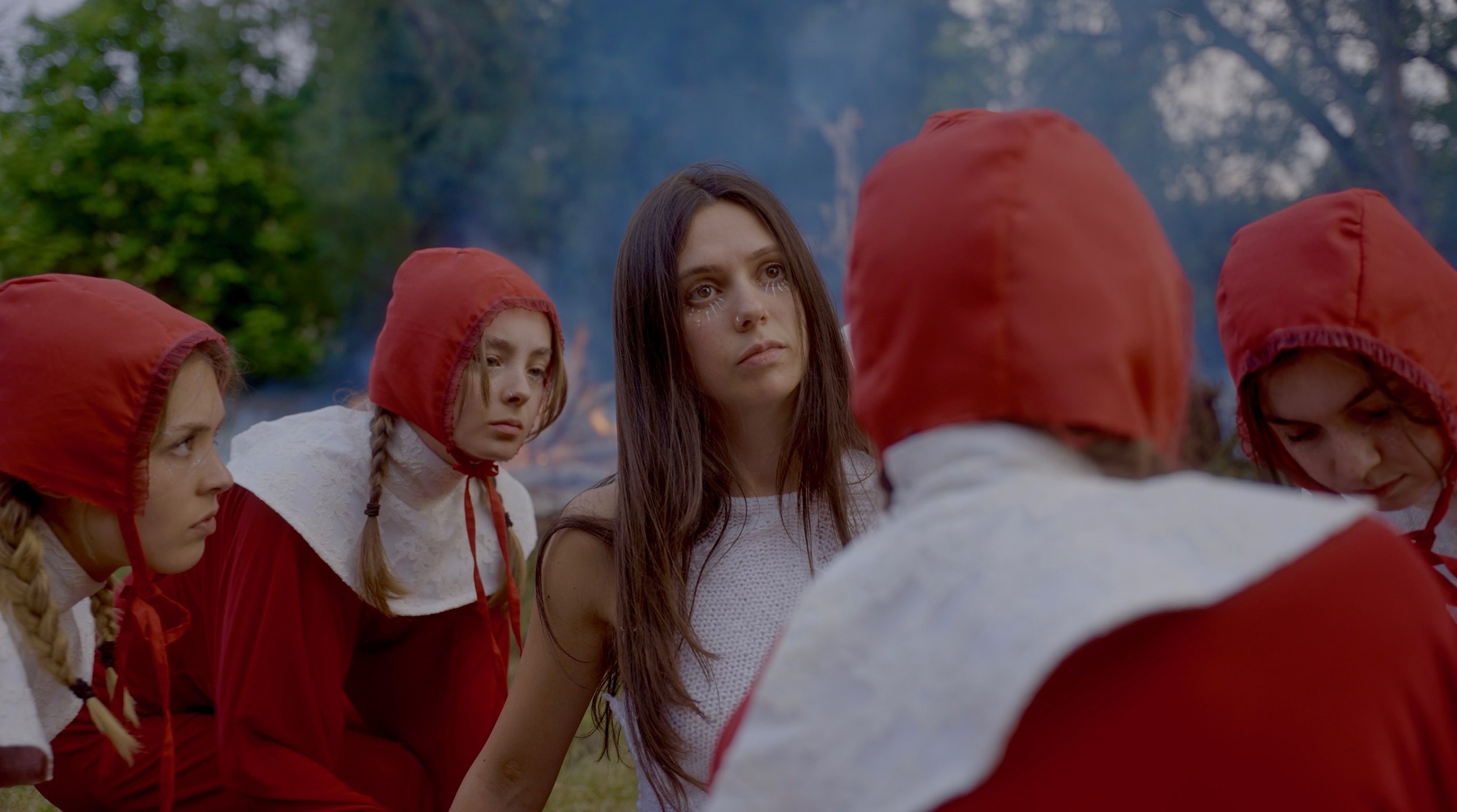 A group of young women, some with braided hair and wearing red hooded cloaks or costumes, gathered in a circle outdoors. One woman with long brown hair and makeup that looks like tears stands in the center, looking serious and contemplative.