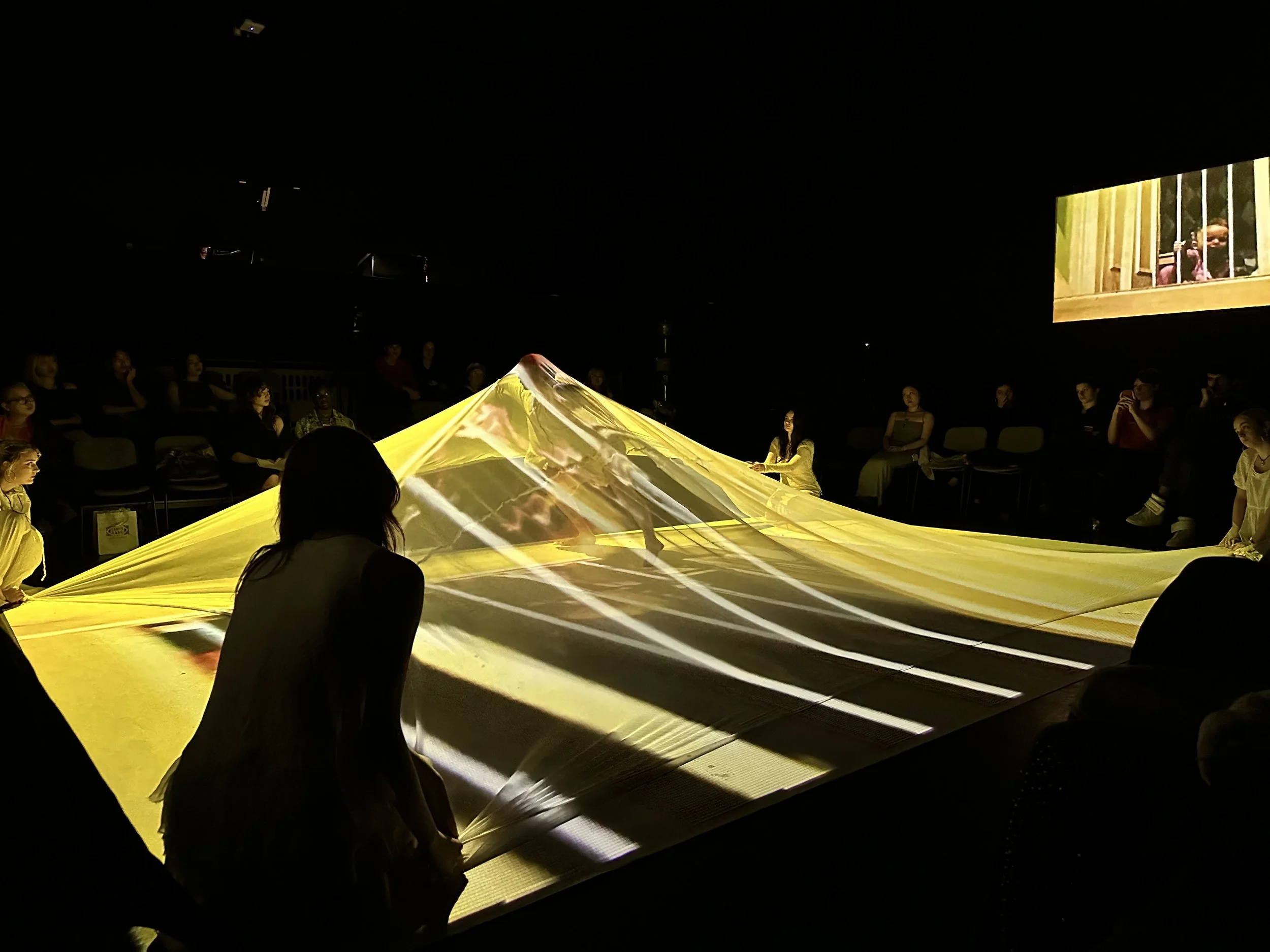 People sitting in a dark room watching a performance involving a large yellow fabric projection and a video screen showing children behind bars.