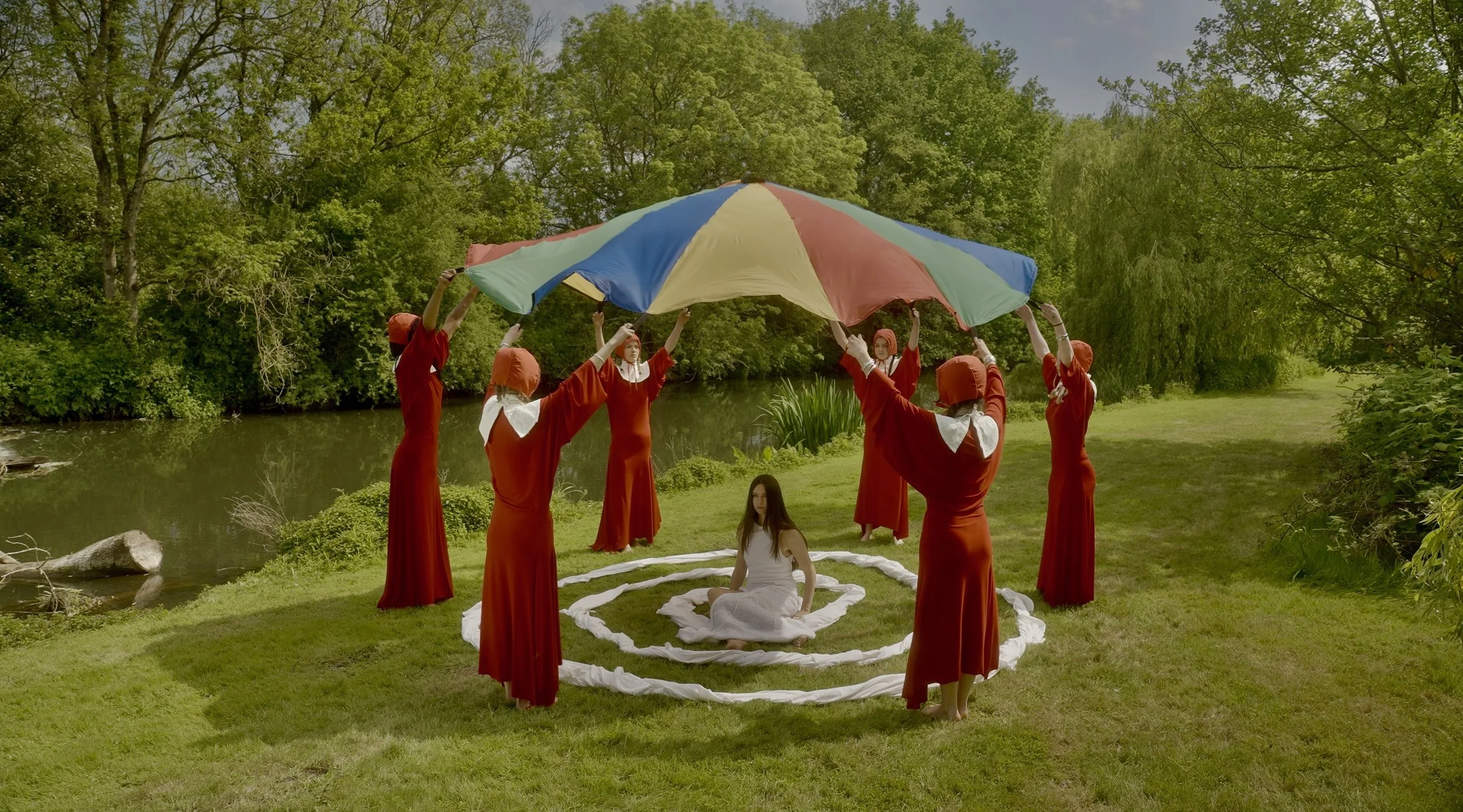 A group of women dressed in red robes and orange head coverings holding a large multicolored umbrella over a woman sitting on the grass in a circular formation near a river, surrounded by lush green trees.
