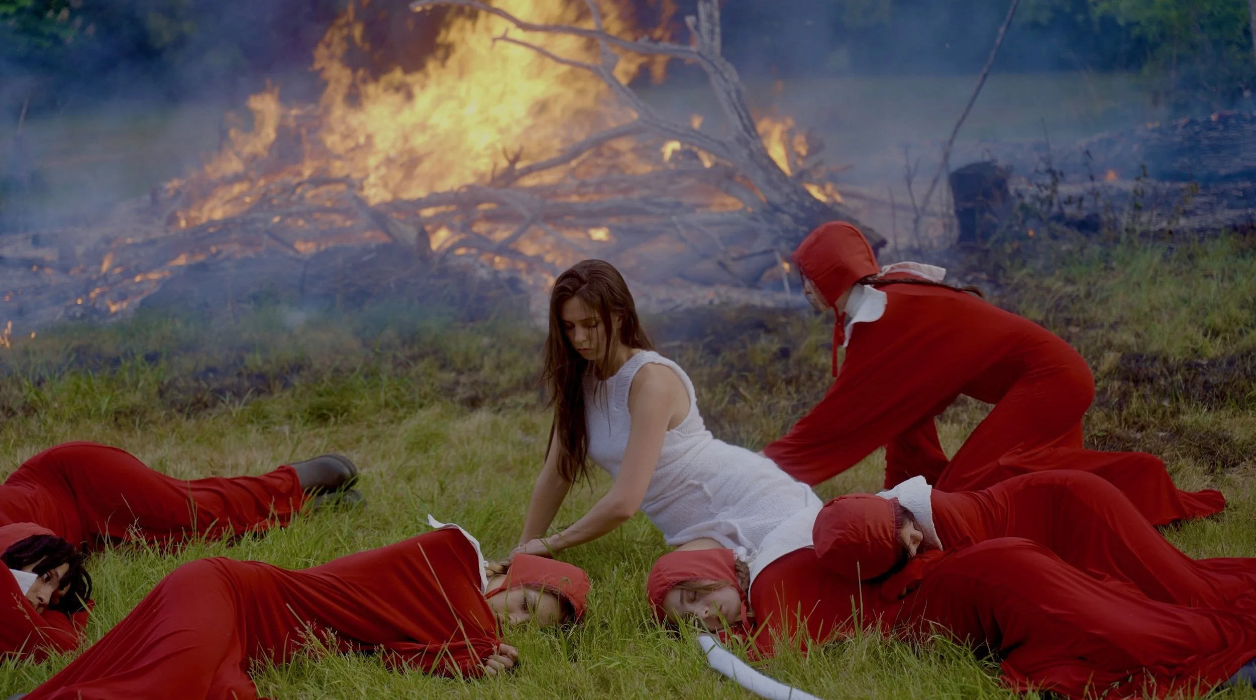 A scene from a movie showing a woman sitting on the grass surrounded by men in red uniforms, with a large forest fire in the background.