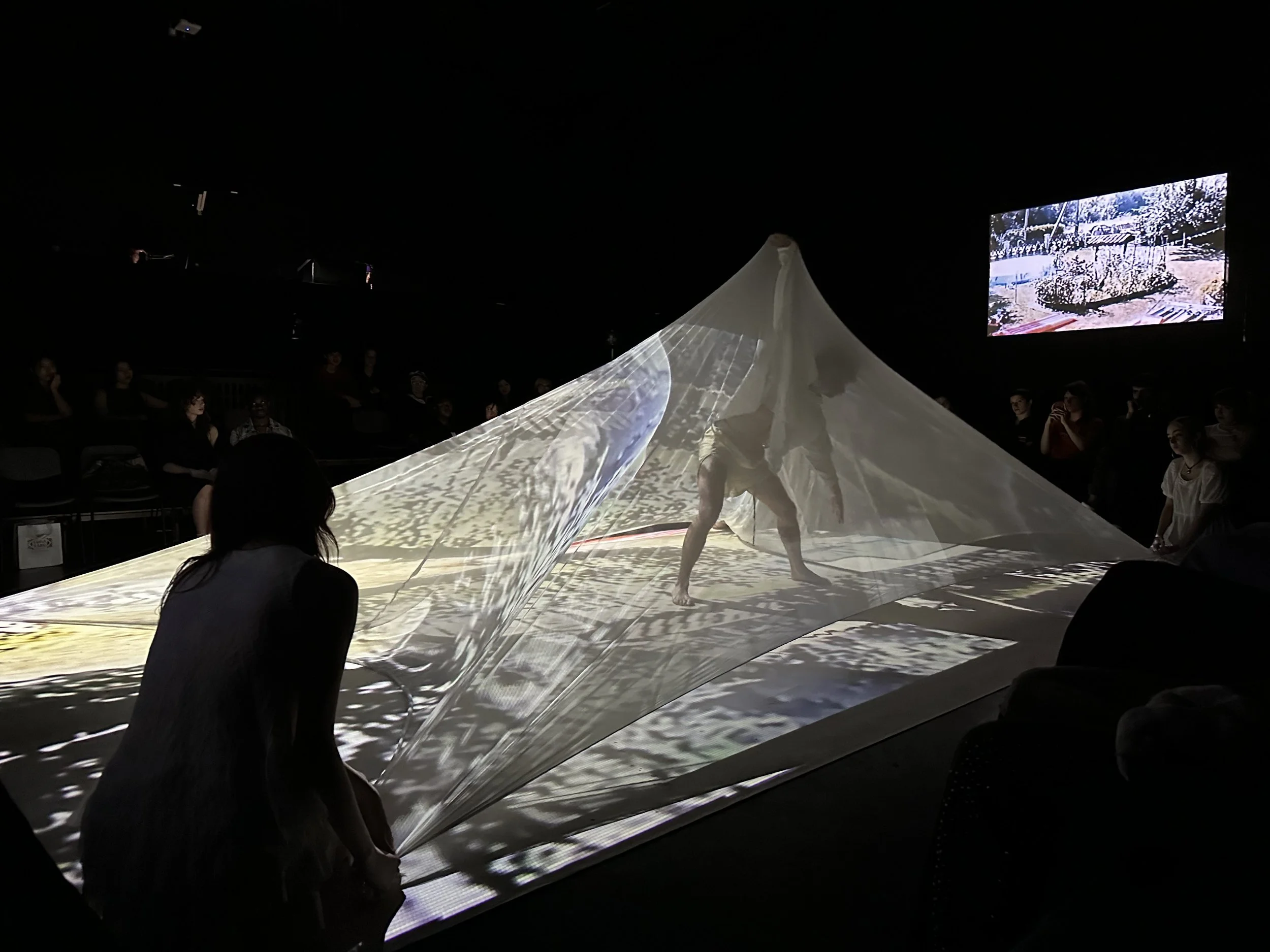 A performance art piece involving a person inside a large, illuminated fabric tent that creates shadows and patterns on the floor, with audience members seated around watching.
