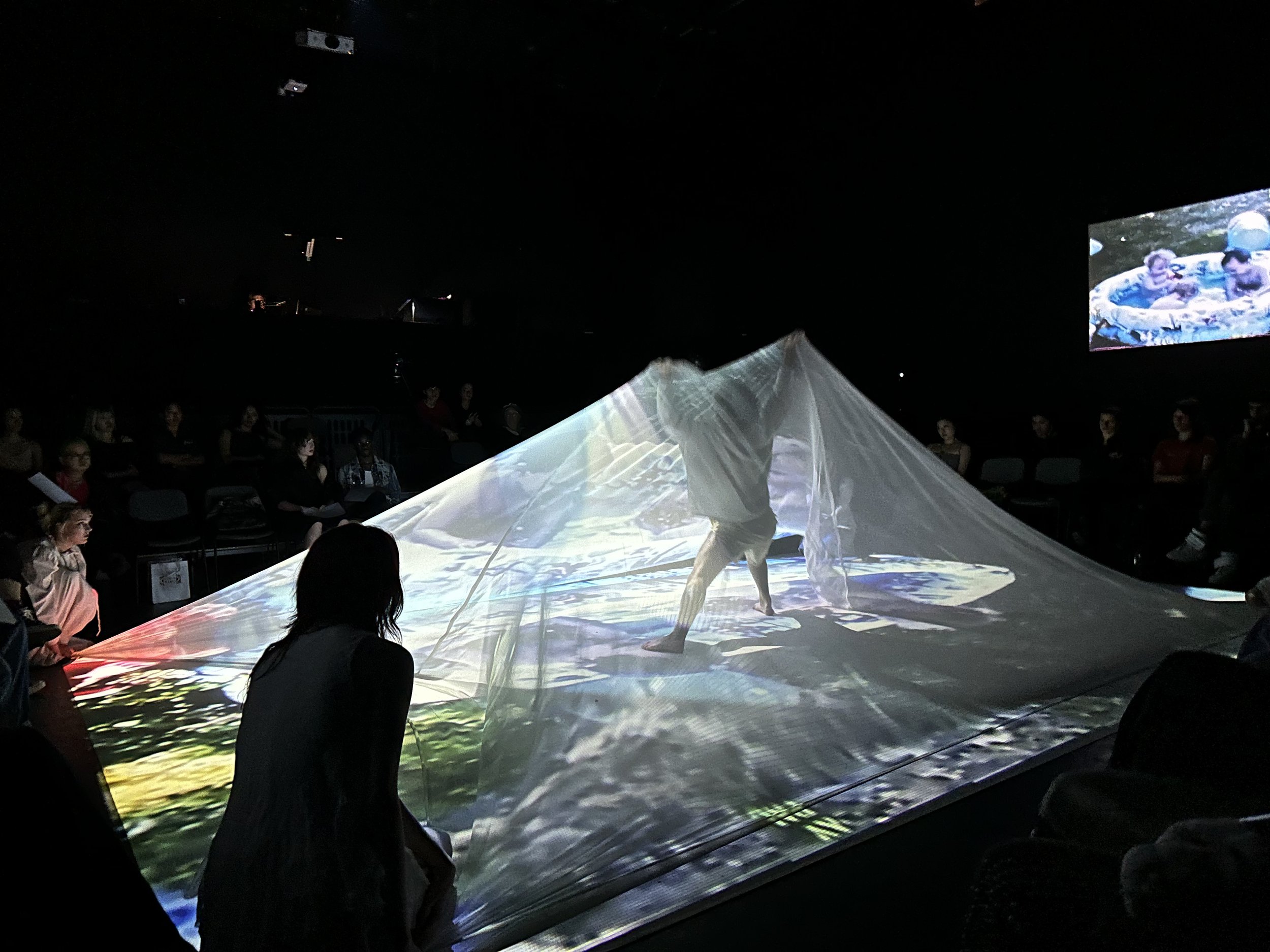 People sitting in a dark room watching a large projection of a scene with two children in a small pool, with a person performing a dance or movement art in the foreground.