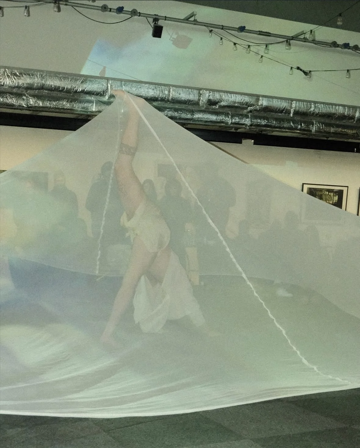 A person performing a dance move on a semi-transparent surface in an art gallery, with other people observing in the background. The surface reflects the person's pose, and the gallery has exposed ductwork and hanging string lights.