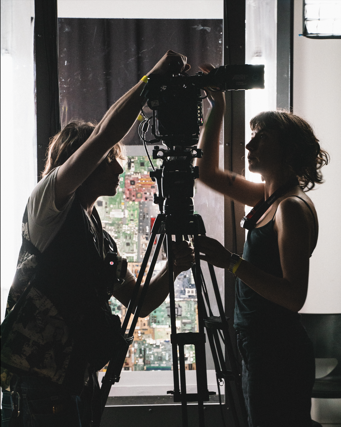 Two women are operating a professional camera on a tripod, silhouetted against a large window showing a cityscape with numerous buildings and streets.