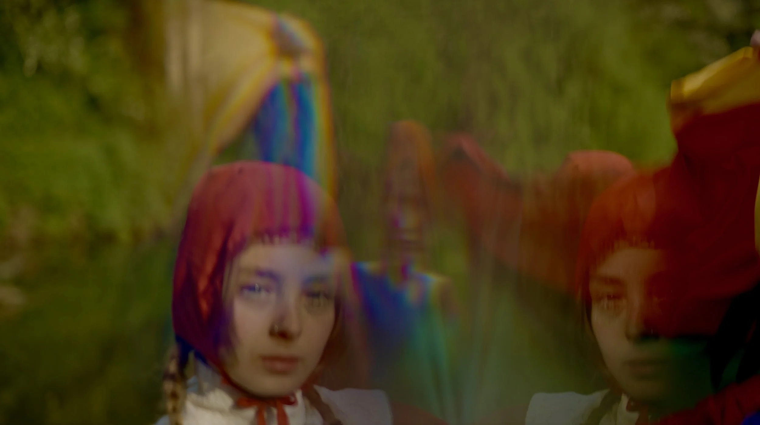 A woman with a rainbow-colored hijab and makeup standing outdoors with green foliage in the background.