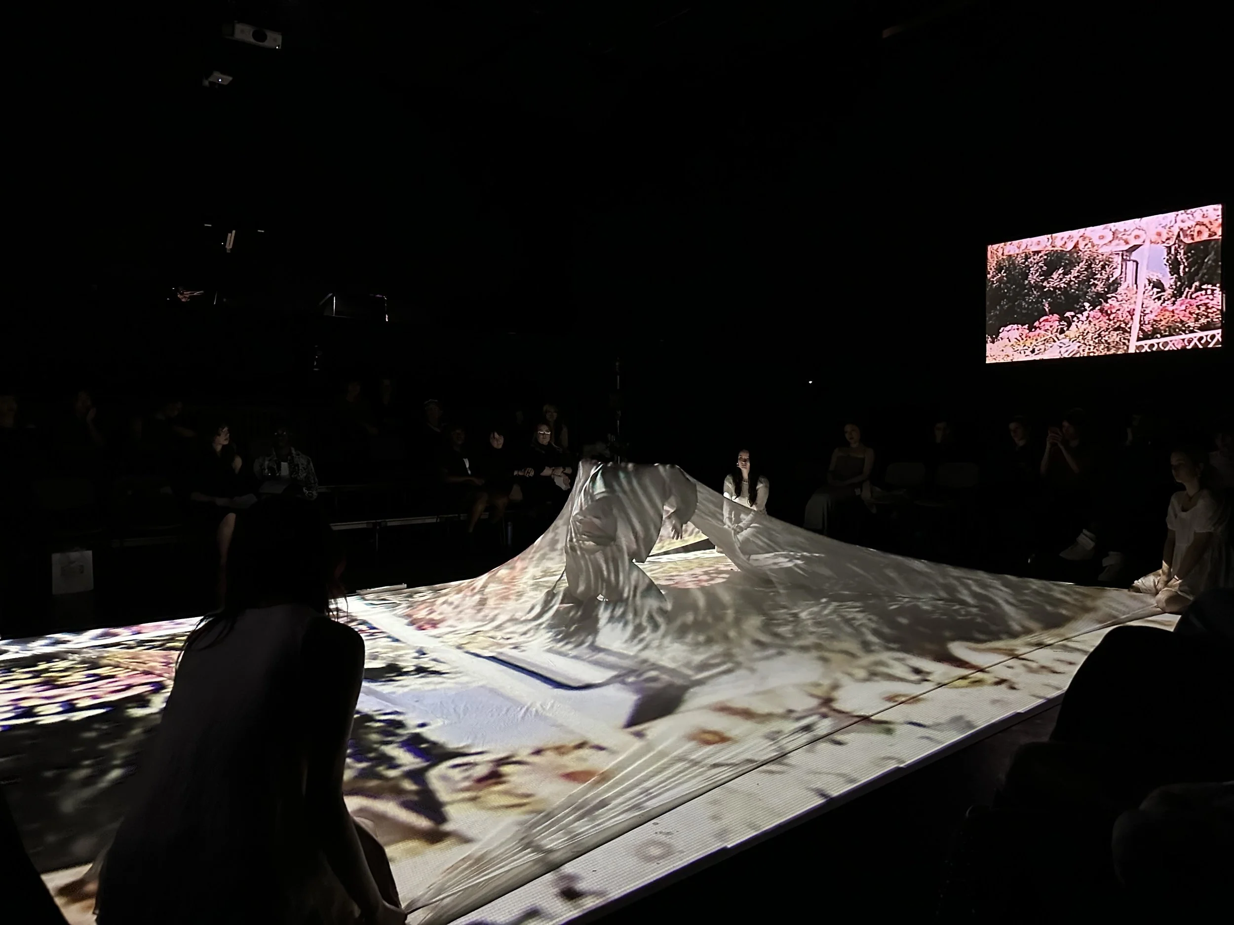 Dark room with projector screens showing pink floral images, audience seated around a table, and a performer in white engaging with a digital or visual art installation.
