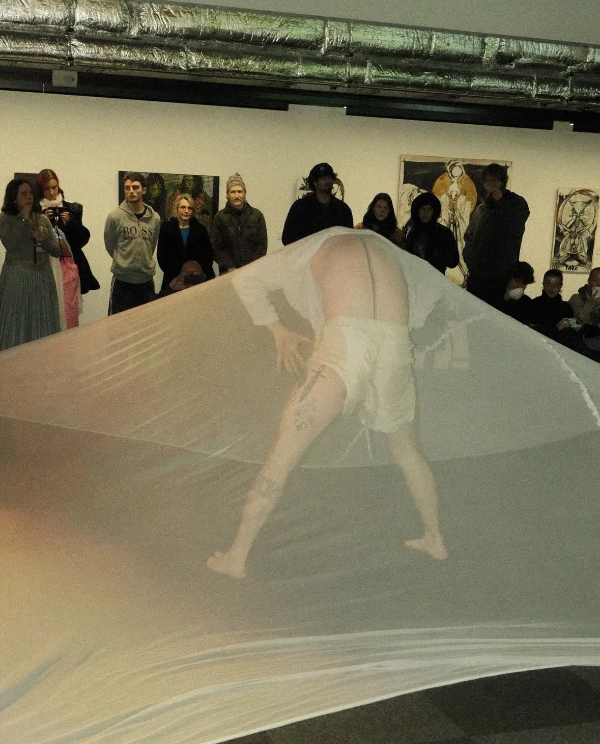 An art exhibition featuring a performance art piece with a person bending forward, partially covered by a transparent sheet or fabric, and tattoos visible on their legs. In the background, a group of people are observing the art, some taking photos, with artwork displayed on the gallery walls.