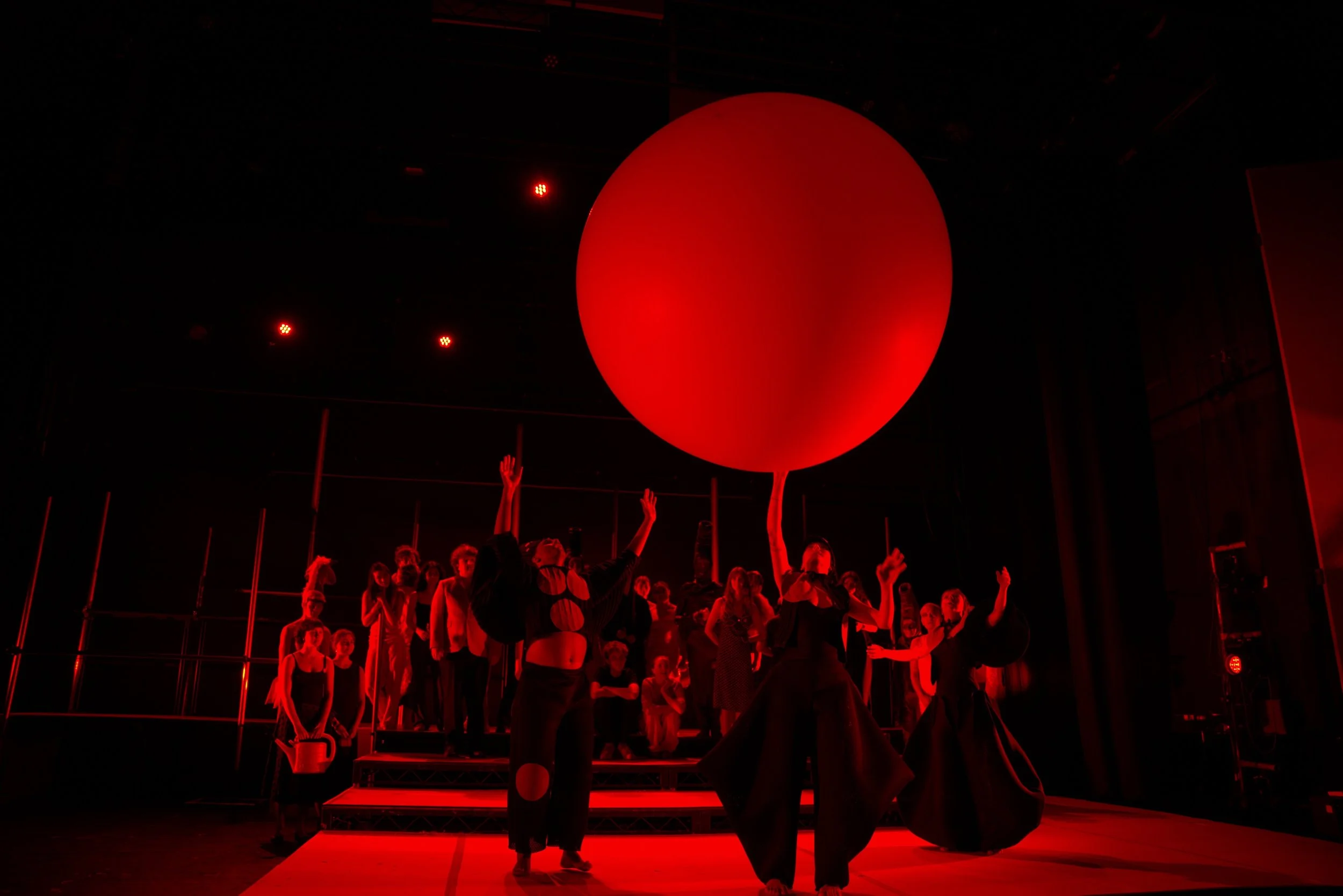 A group of performers on stage with a large red balloon, all dressed in black, illuminated by red lighting, during a theatrical performance.