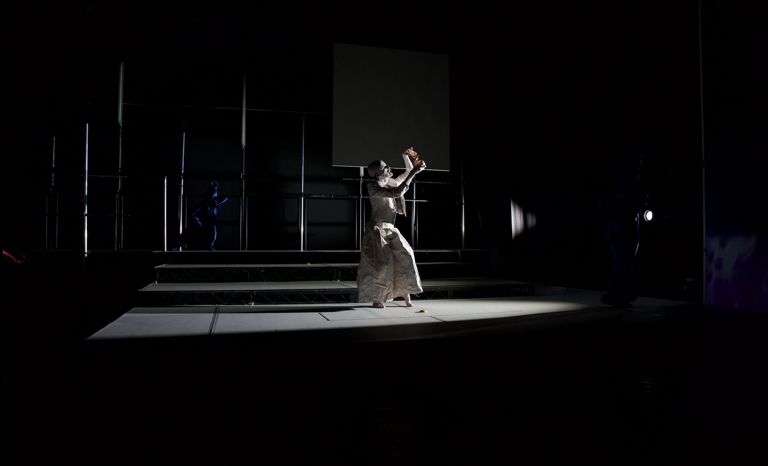 Performer dressed as a mime with white face paint, black and white clothing, in a theatrical scene on stage with minimal lighting. The mime appears to be holding a puppet or object above their head.