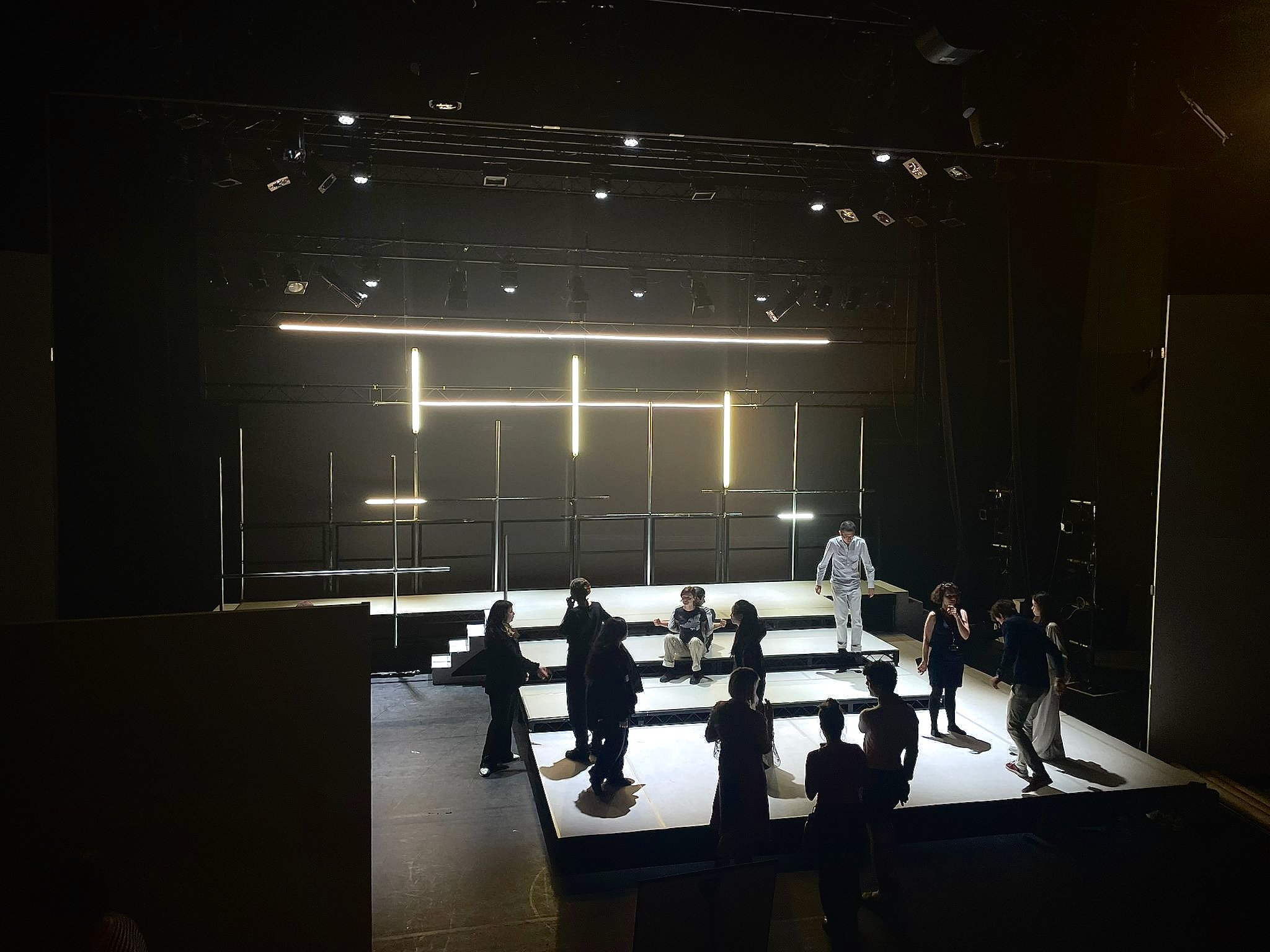 A dark theater stage with minimalistic lighting and several people setting up or rehearsing. The stage has illuminated horizontal and vertical lines, with some people standing and talking near the front. The background is mostly black.