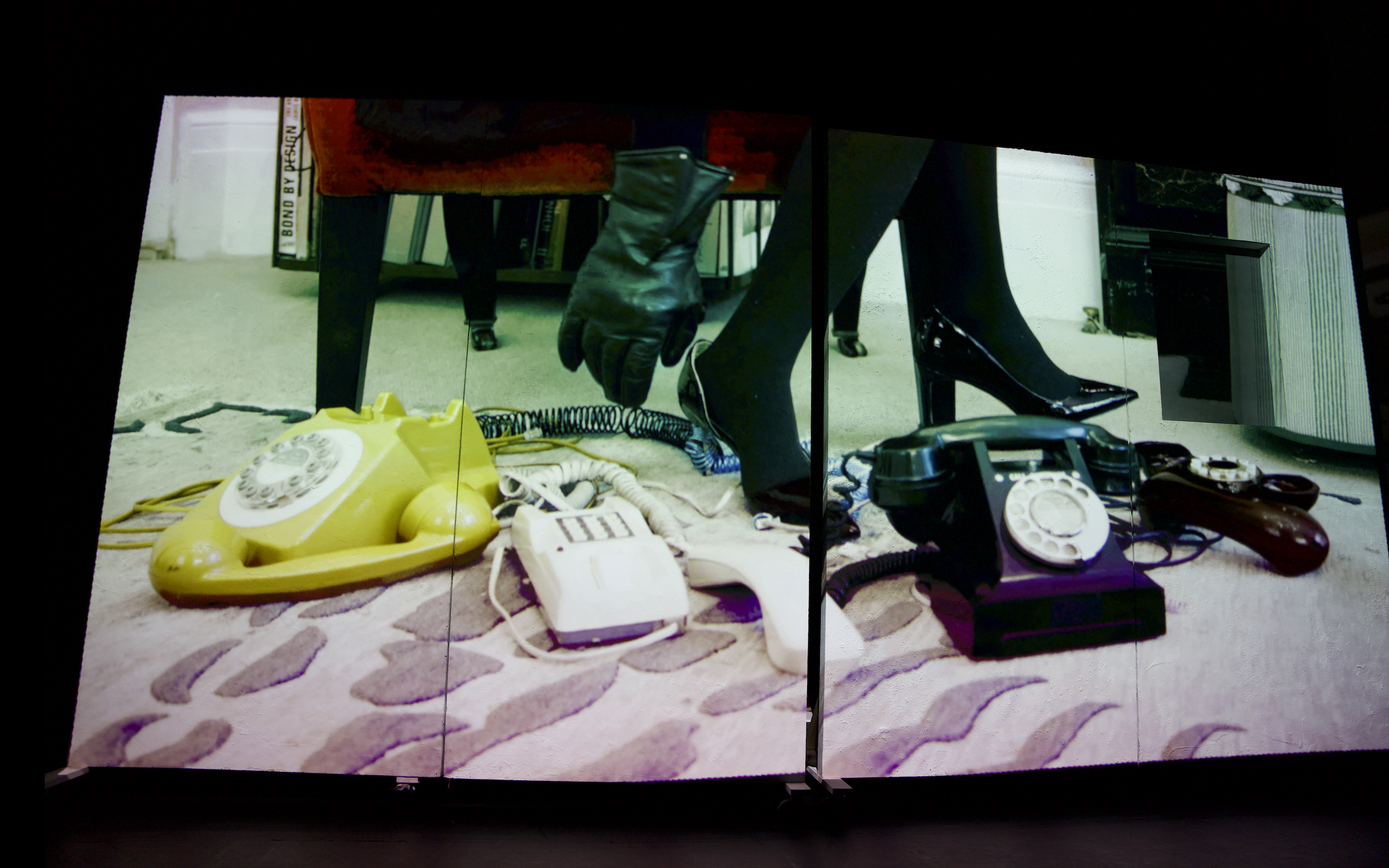 Display of vintage objects including a yellow rotary phone, an old white remote control, and black wired telephone receivers on a patterned carpet, viewed through a reflective surface.