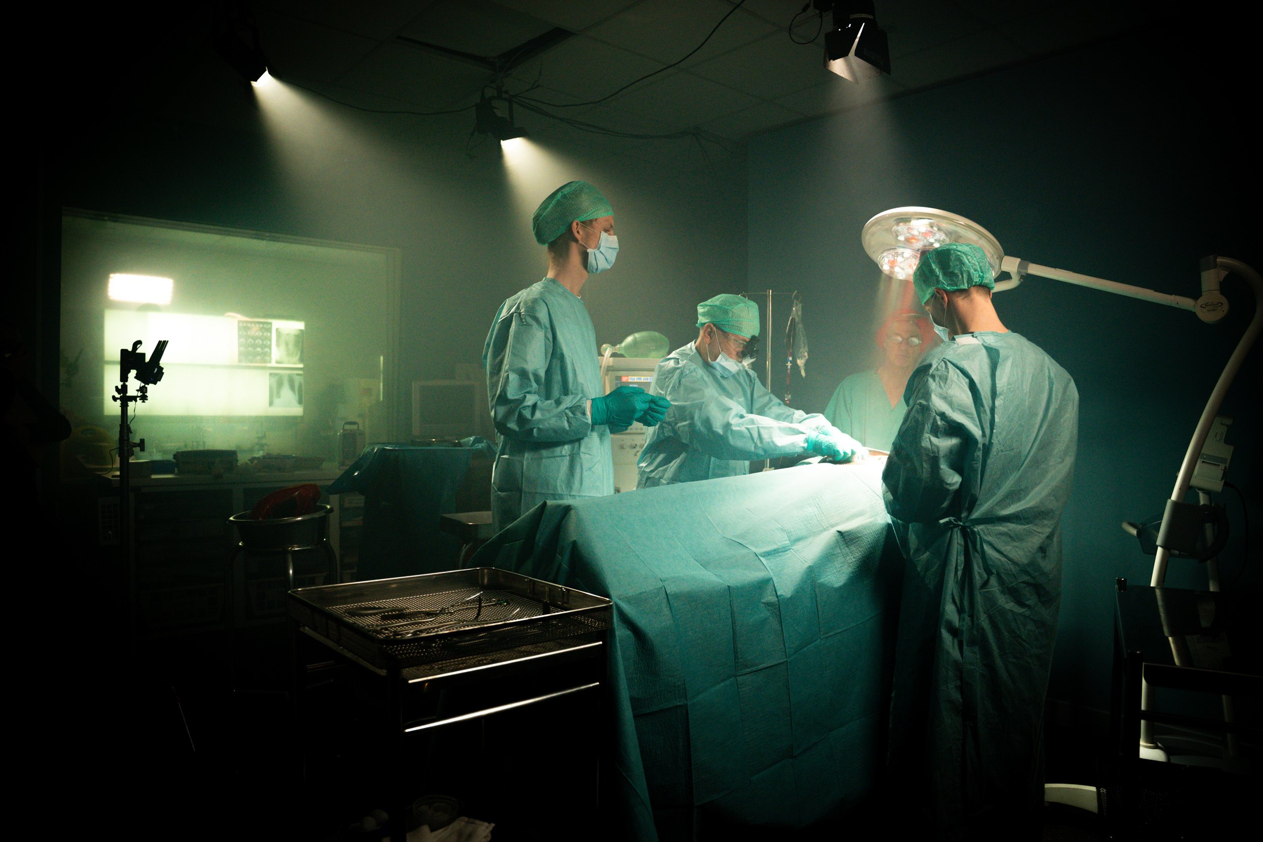 Medical team performing surgery in an operating room with surgical lights overhead, healthcare professionals wearing scrubs, masks, and caps, and medical equipment around them.