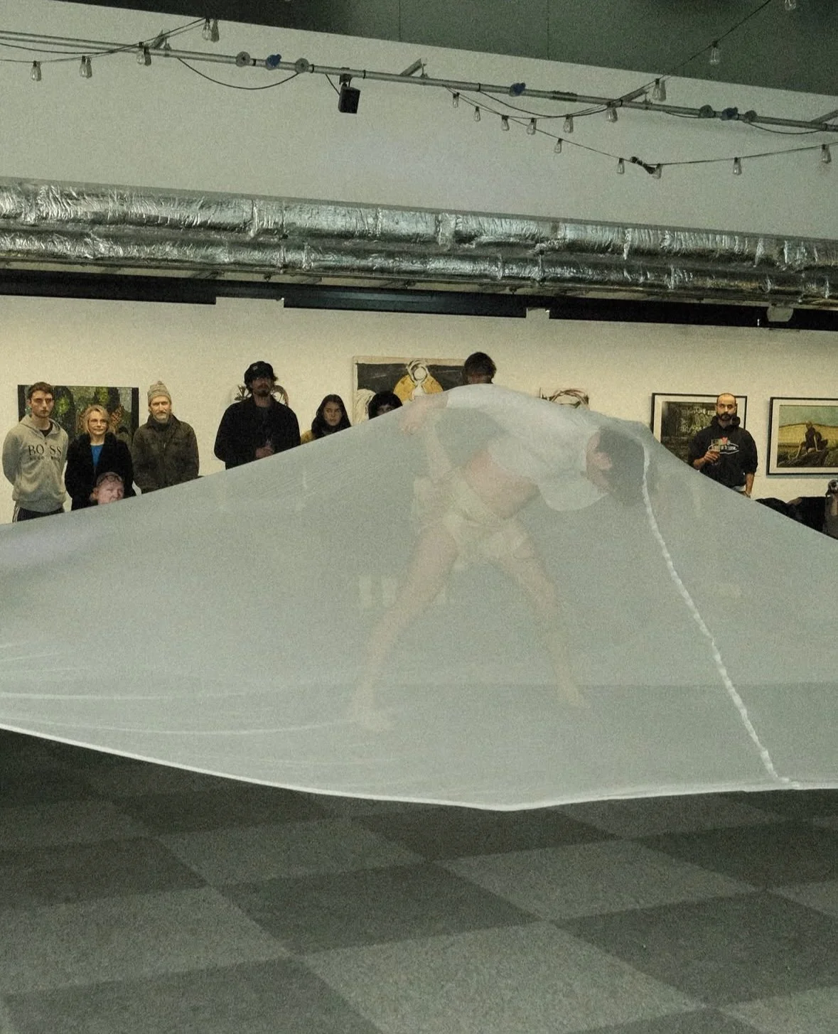 A group of people watching performers on stage, with two performers under a large semi-transparent fabric or parachute, indoors with artwork on the walls and exposed air ducts.
