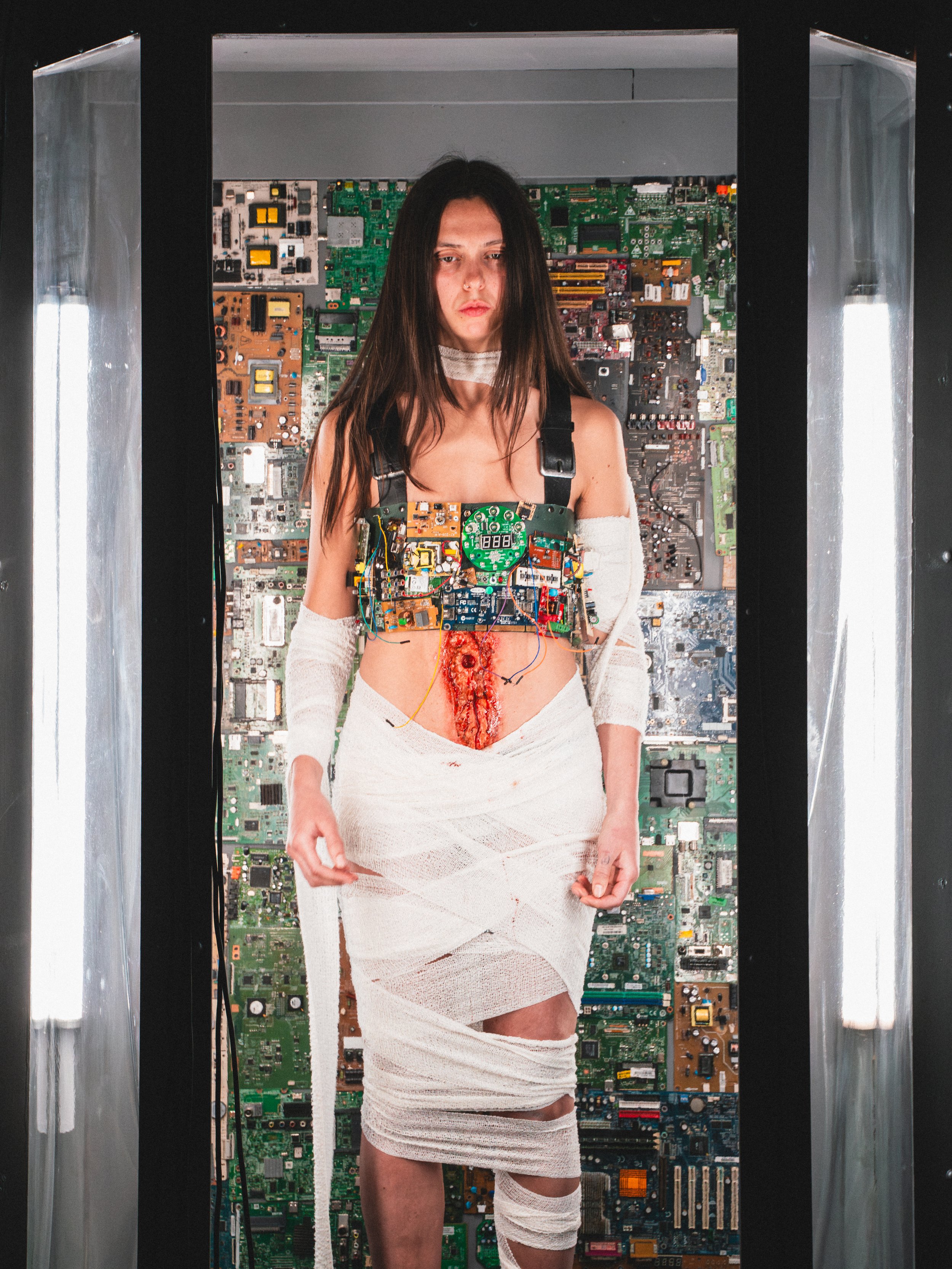 A woman in a white dress with visible wounds and electronics on her torso is standing inside a futuristic elevator with walls covered in circuit boards.