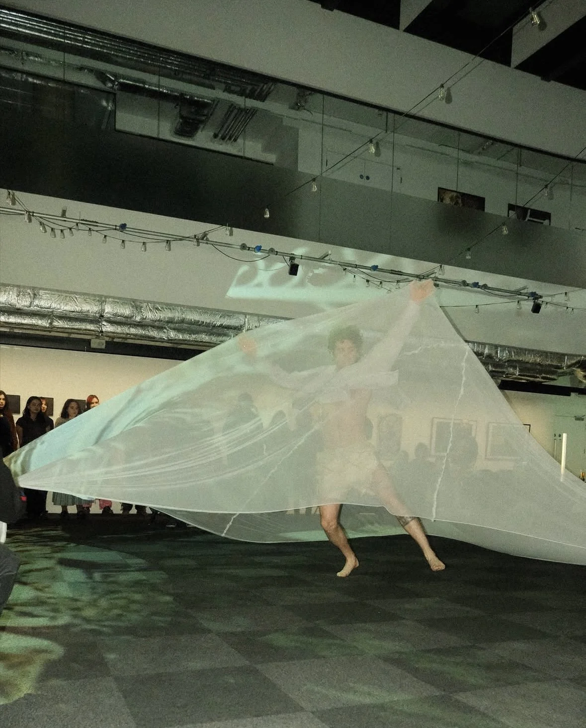 A performer dancing behind a large, flowing, semi-transparent fabric in an art gallery or exhibition space. Several onlookers are observing the performance, with artwork visible on the walls.