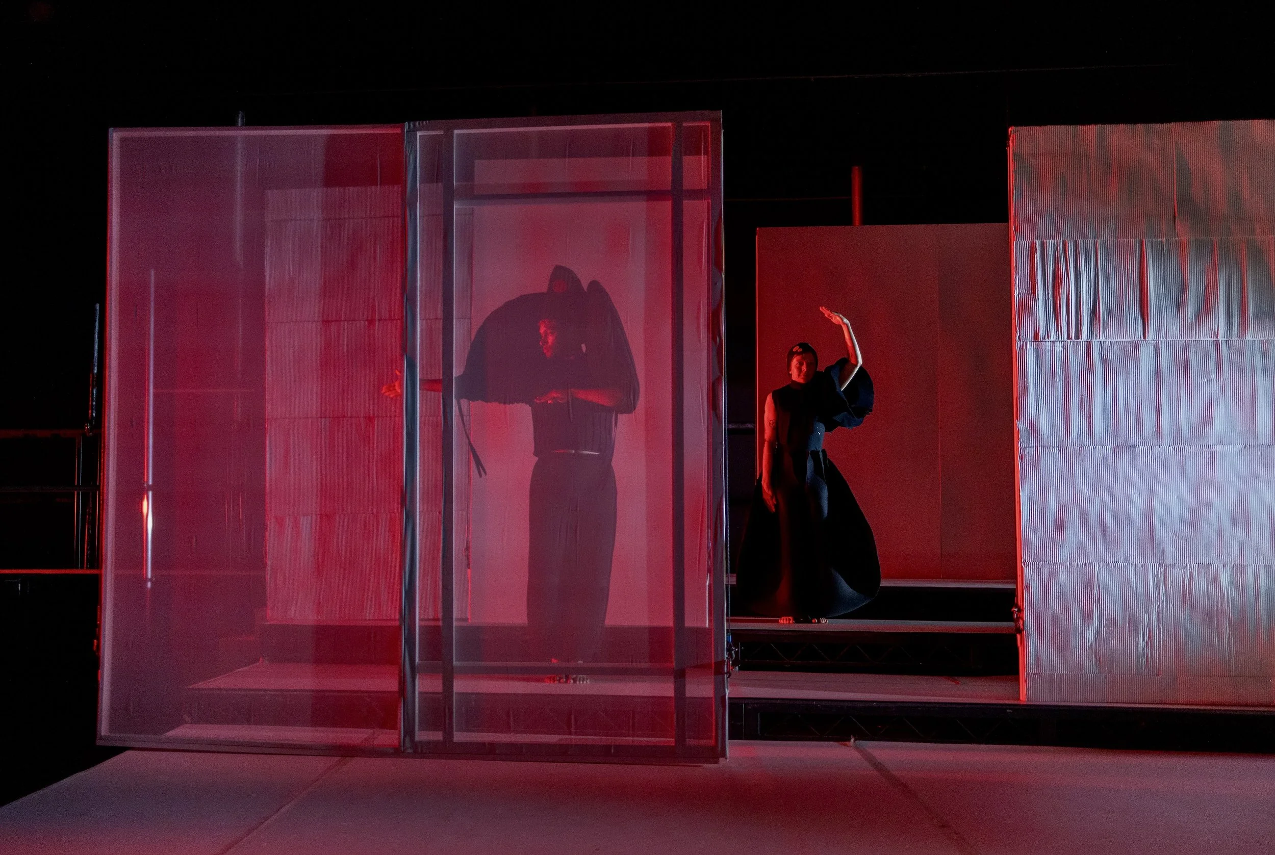 A person with a raised arm standing next to a transparent glass display case illuminated with red lighting, with black background and textured panels.