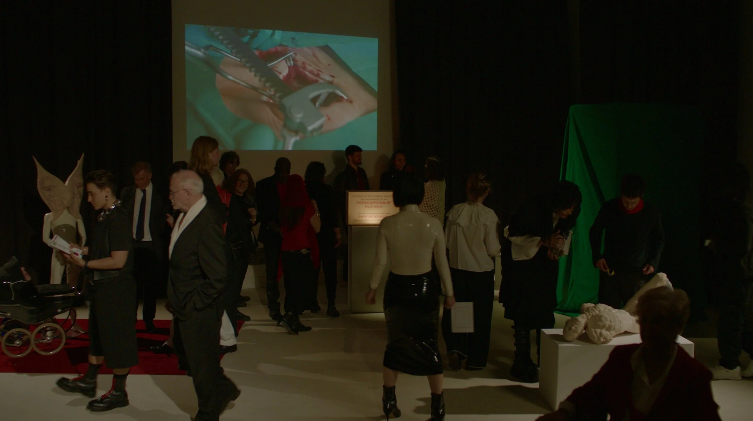People gathered in a dark room, some looking at sculptures, with a large screen displaying an image of surgery involving a hand and medical tools.
