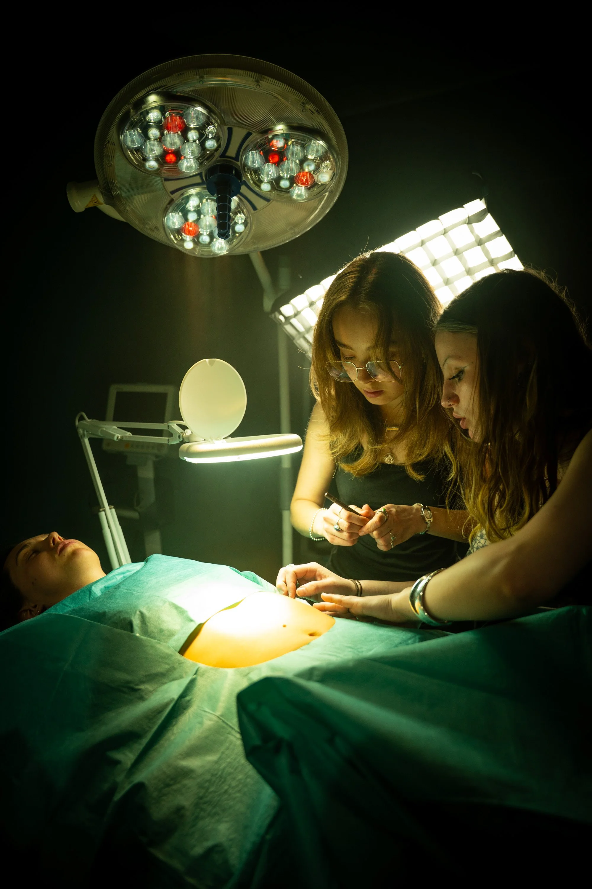 Medical emergency room scene with two women attending to a patient lying on a hospital bed, under surgical lights.