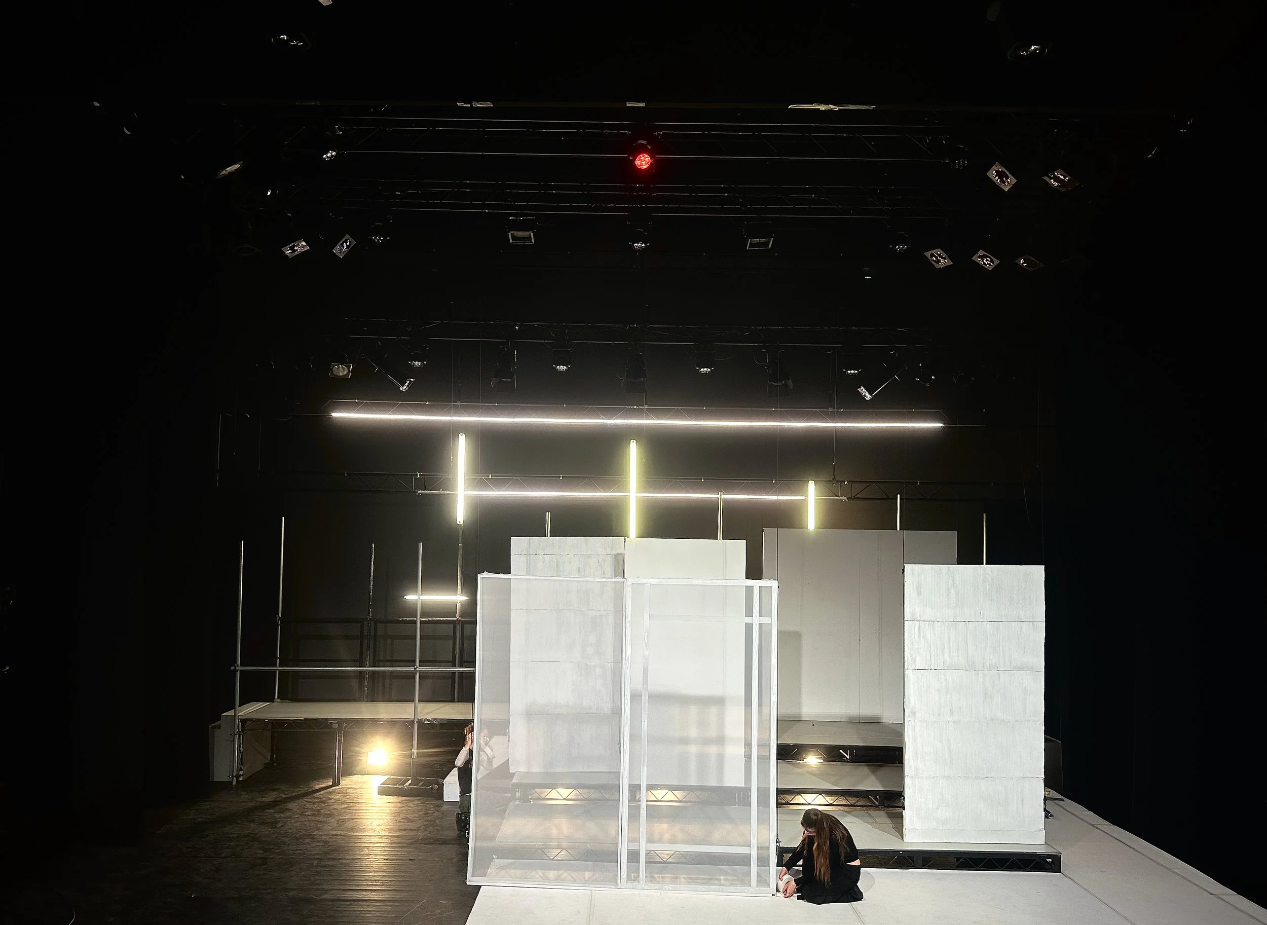 Stage set with scaffolding, white panels, and minimal lighting, with two individuals working on the setup.