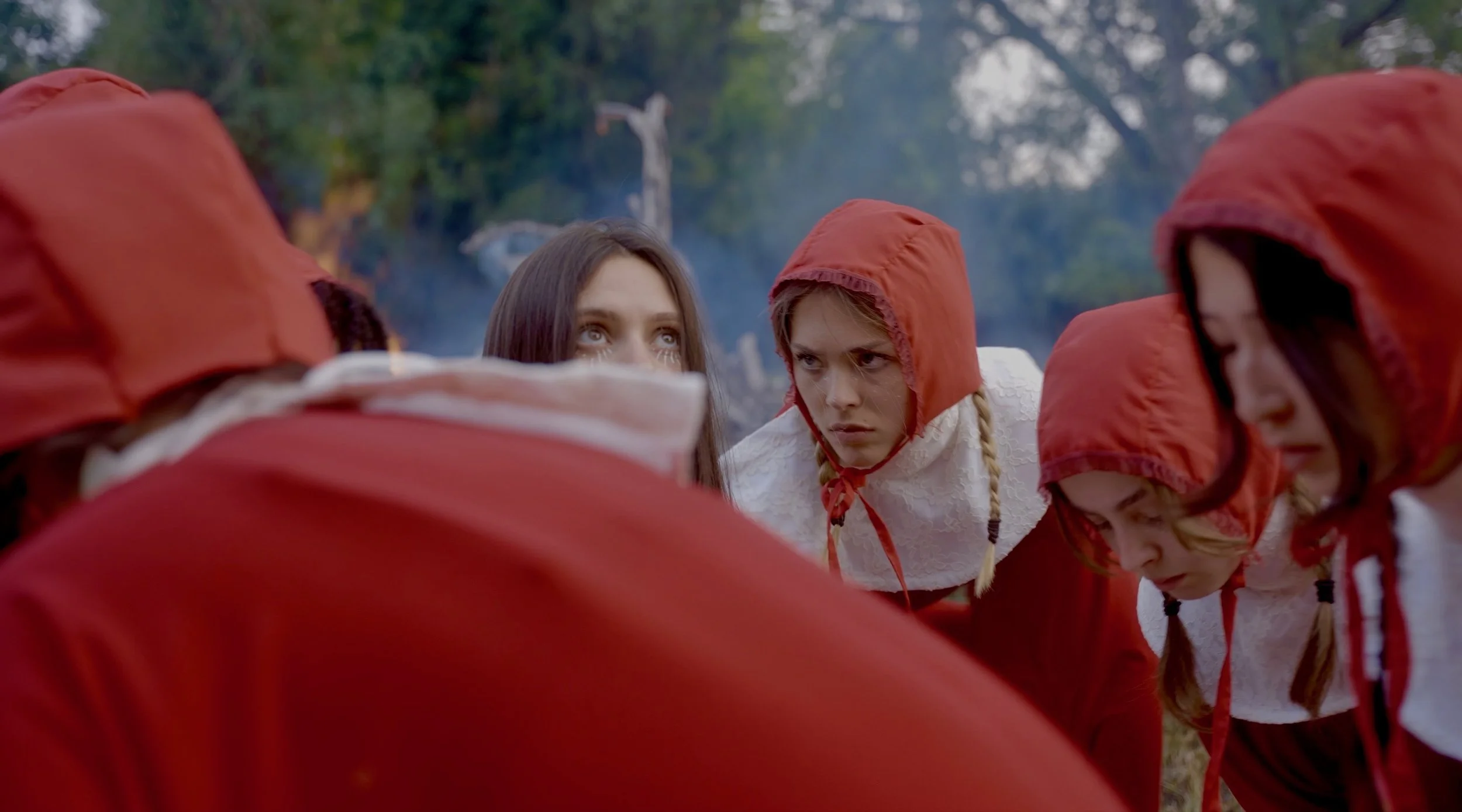 A group of young women and girls dressed in white with red hoods, are in a huddle outdoors, surrounded by trees, with focused expressions.