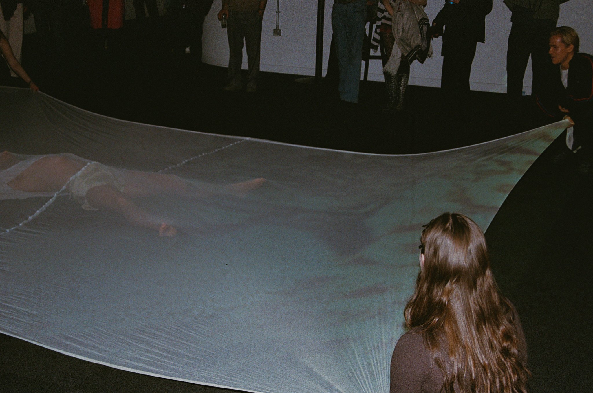 People are gathered in a dark room, some holding a large fabric or sheet, engaging in what appears to be an interactive or theatrical activity.