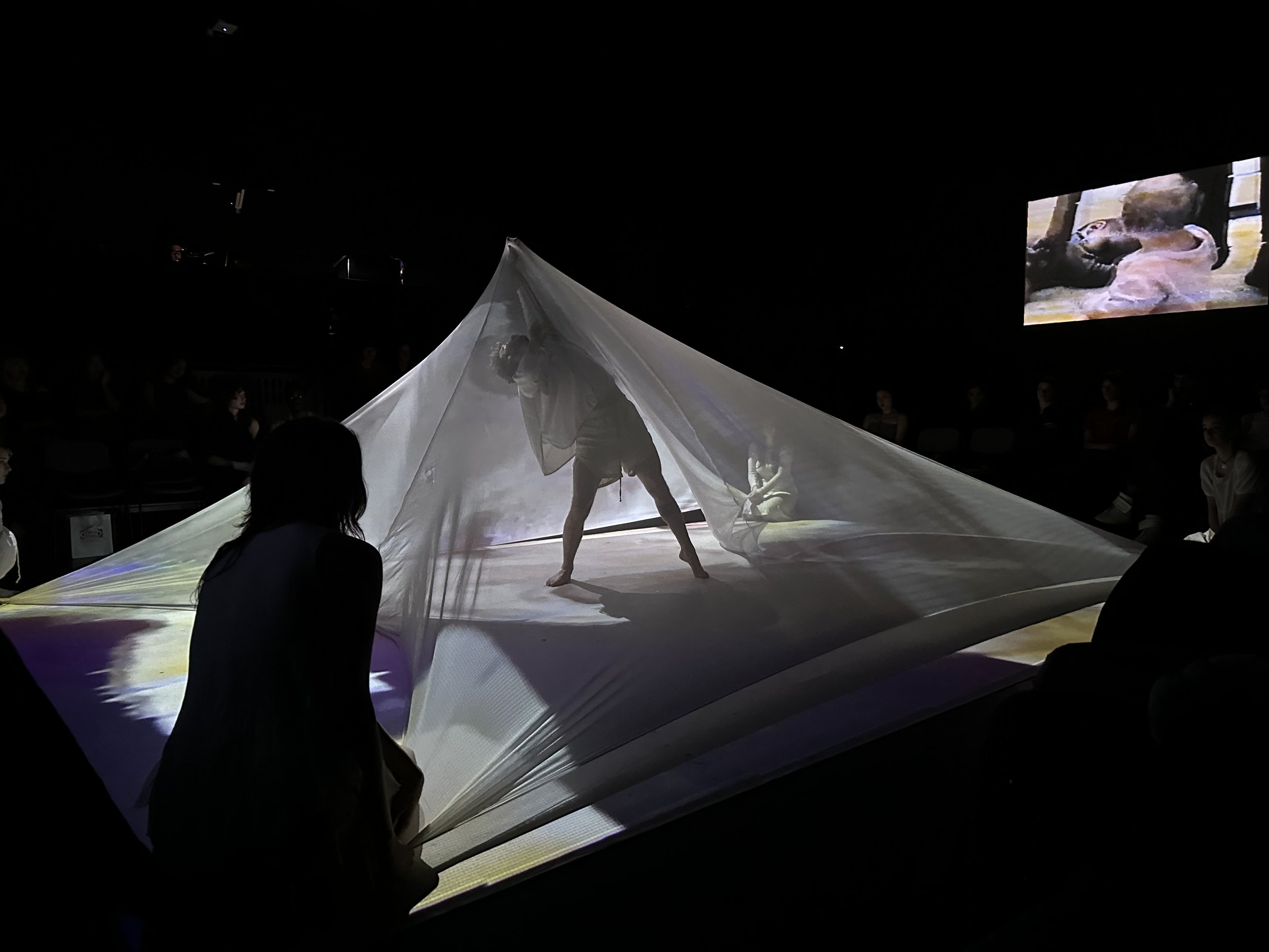 A performer on a stage with a large, billowing white fabric at a live event, with a video screen showing another performer in the background.