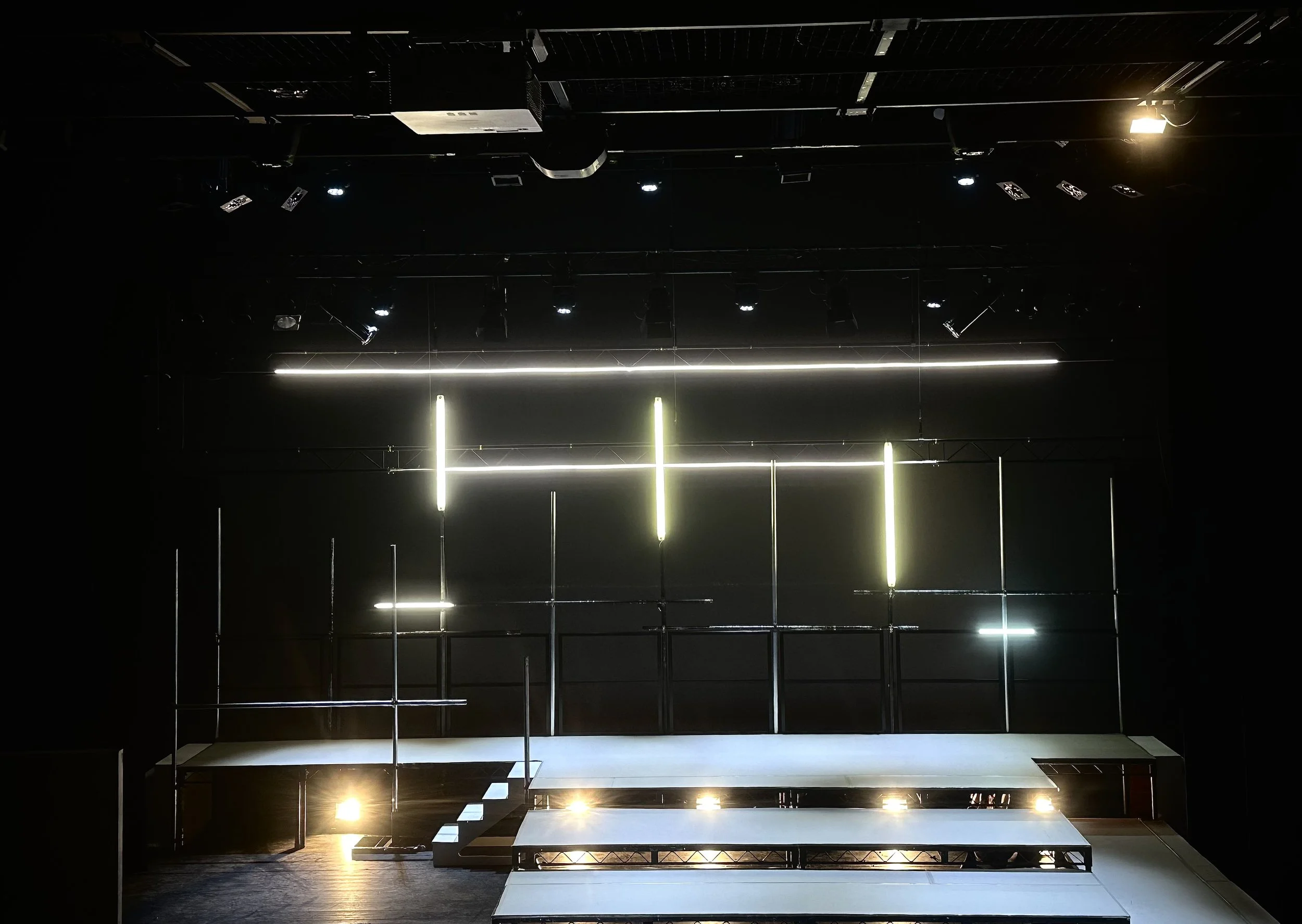 A dark stage with black walls, illuminated by vertical and horizontal light strips. There are spotlights and equipment hanging from the ceiling, and a set of stairs on the left side.