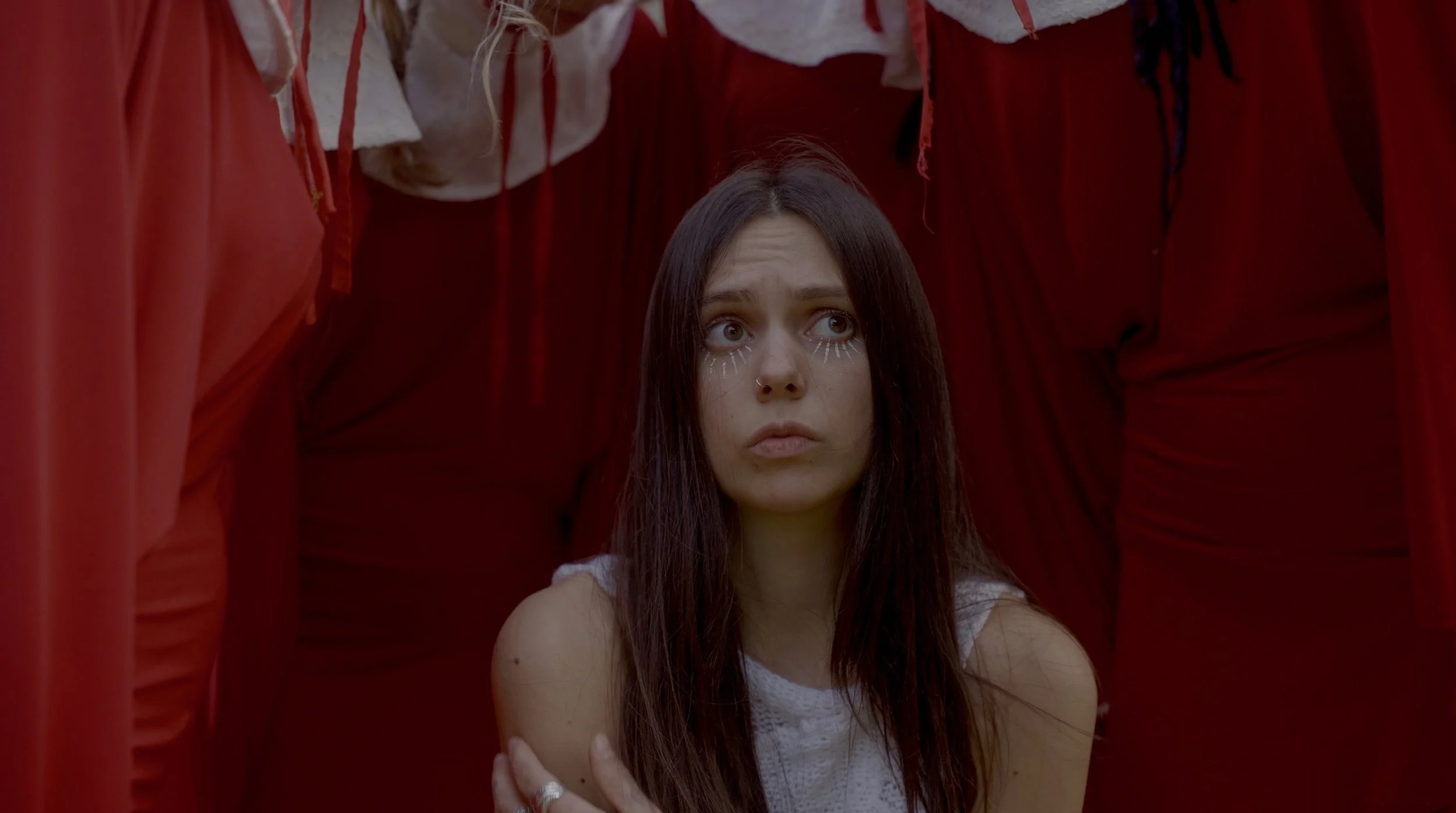 A young girl with long dark hair and white face paint, sitting among people dressed in red robes and hoods.
