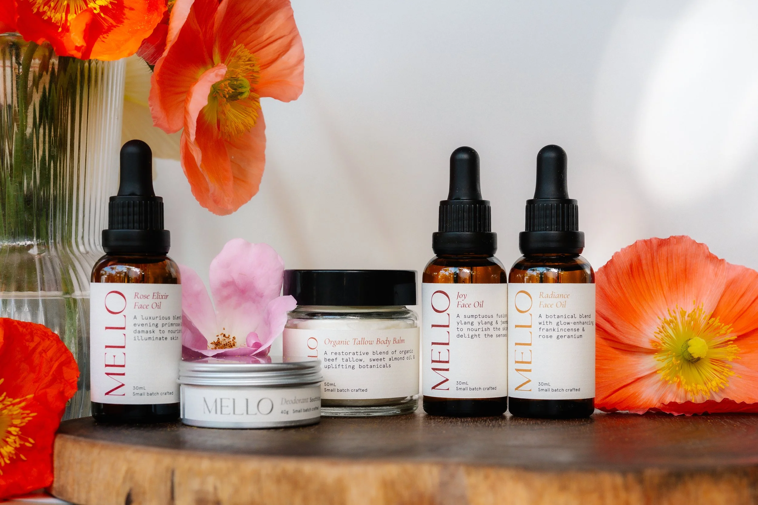 A display of skincare products with white labels, surrounded by vibrant orange and pink flowers, on a wooden surface.