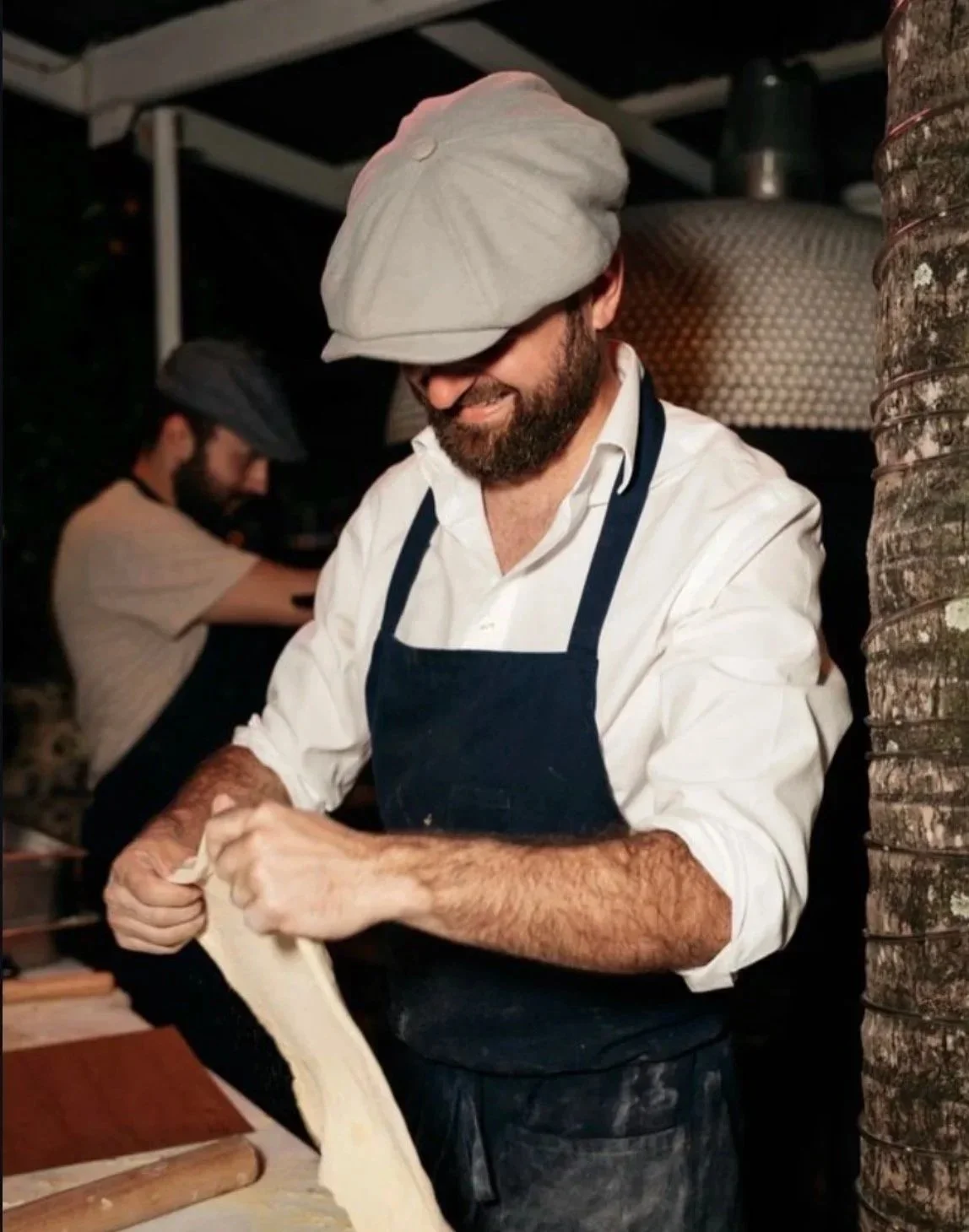 A man wearing a chef's hat and apron is stretching dough, smiling, in a kitchen with another person in the background.