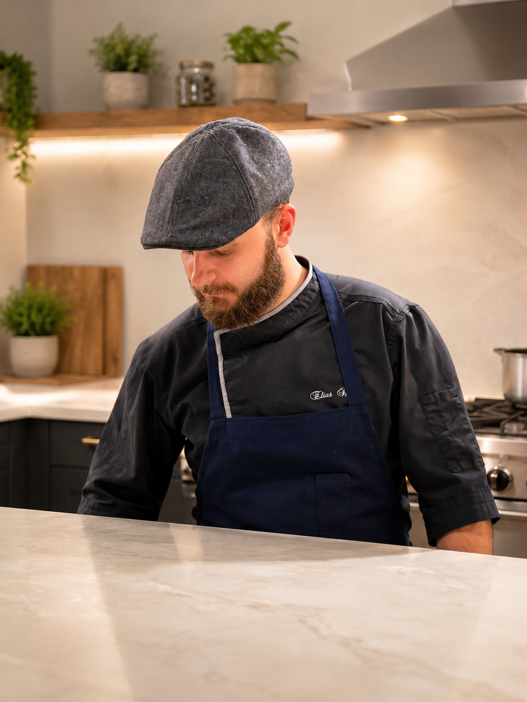 A man in a chef's uniform and a gray cap standing at a kitchen counter, looking down.