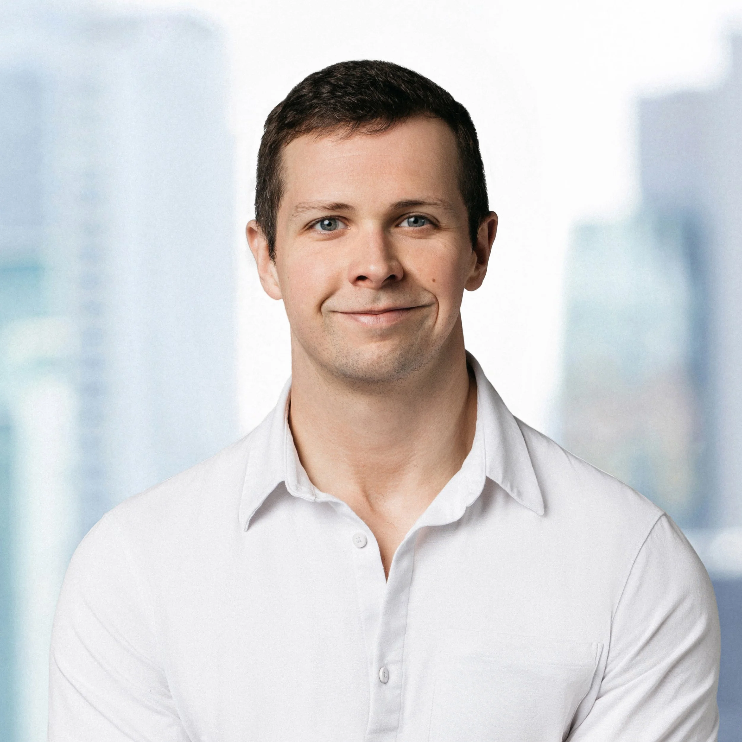 A professional corporate headshot of aClose-up image of a person with short dark hair, blue eyes, wearing a white-collared shirt, smiling slightly, with a blurred light-colored background.