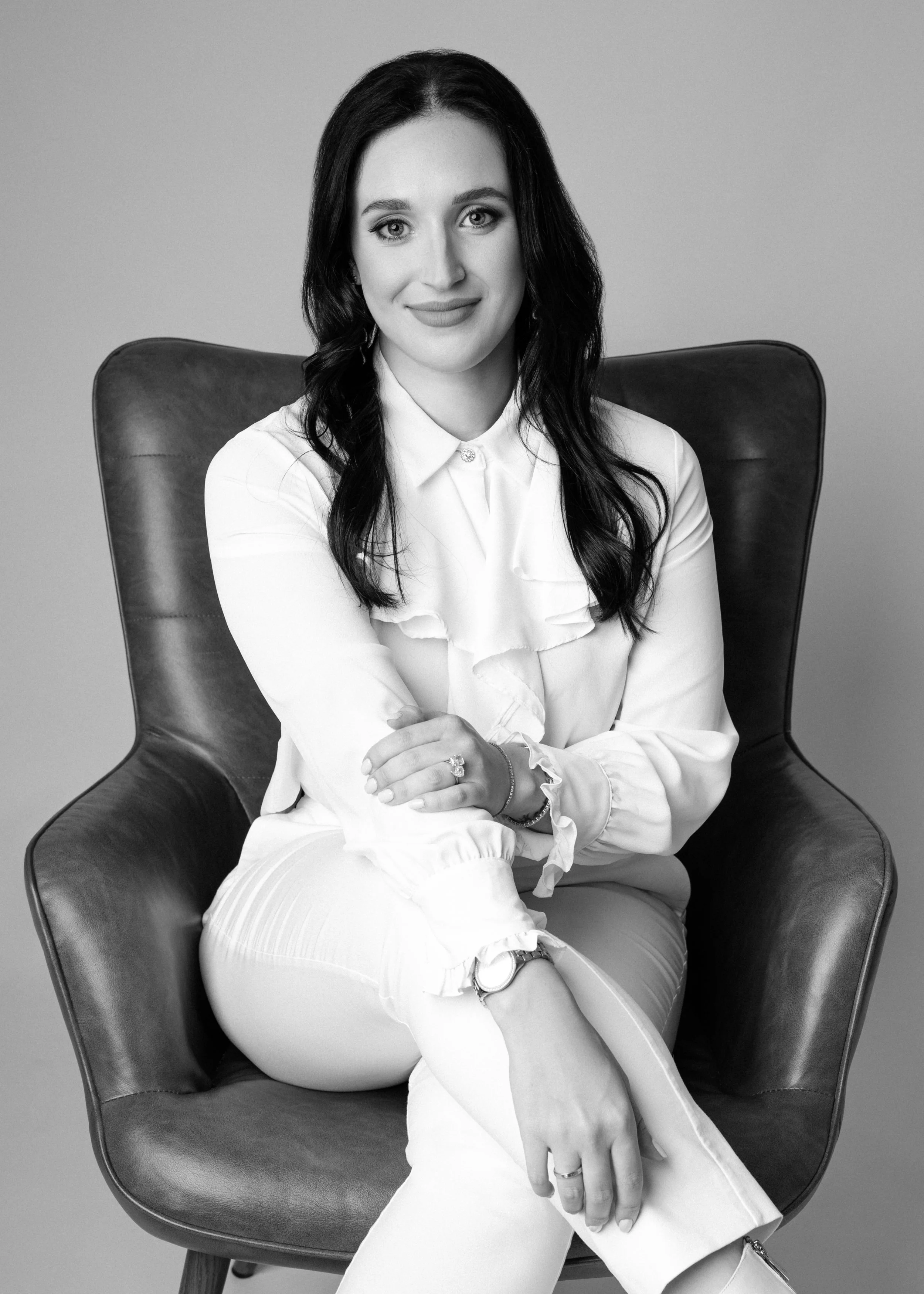 A professional corporate headshot of a woman with long dark hair sitting on a modern armchair, wearing a white blouse and white pants, smiling confidently, with her arms crossed over her lap.